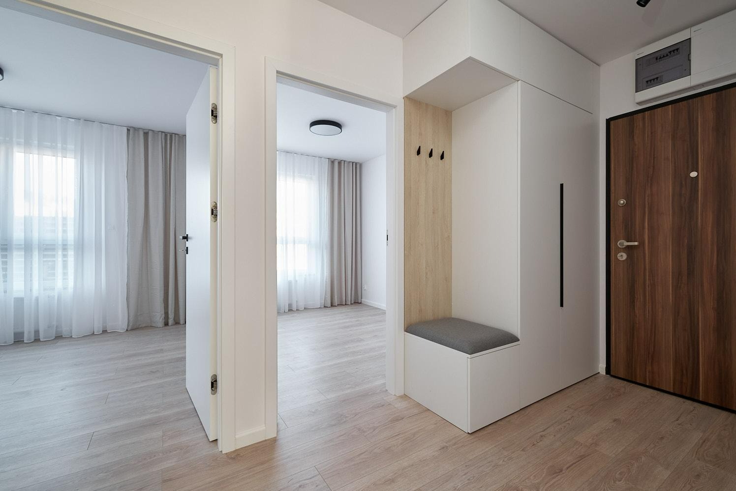 Nowoczesny przedpokój z drewnianą podłogą, białymi ścianami, zabudowaną szafą z siedziskiem i dwiema otwartymi przestrzeniami prowadzącymi do innych pomieszczeń, widoczne drzwi wejściowe z ciemnego...