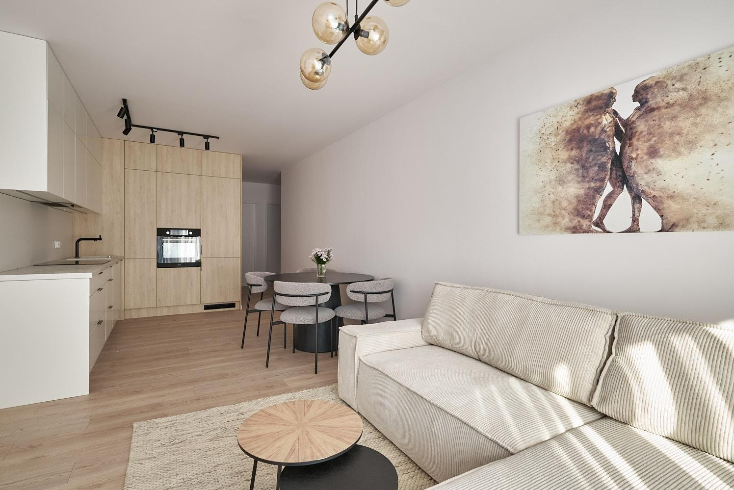 Jasne wnętrze mieszkania z aneksem kuchennym, okrągłym stołem z krzesłami i beżową sofą, oświetlone lampą sufitową z kulistymi kloszami i ozdobione obrazem na ścianie.