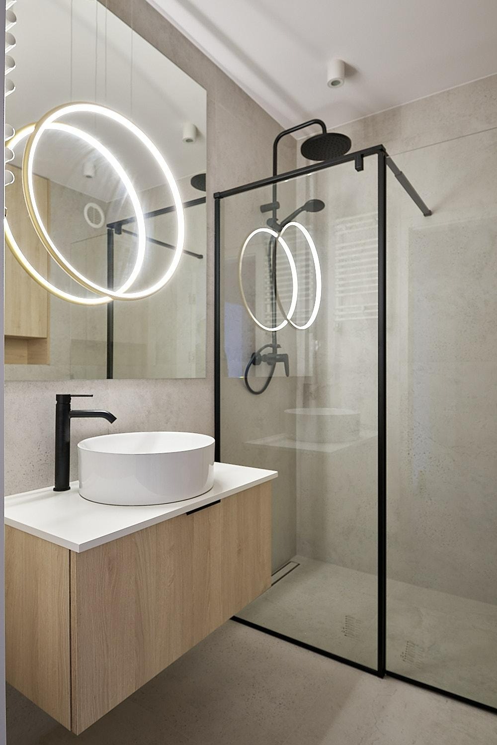 Nowoczesna łazienka z okrągłą umywalką nablatową, czarną armaturą i kabiną prysznicową typu walk-in z czarnymi profilami, oświetlona lampą pierścieniową.