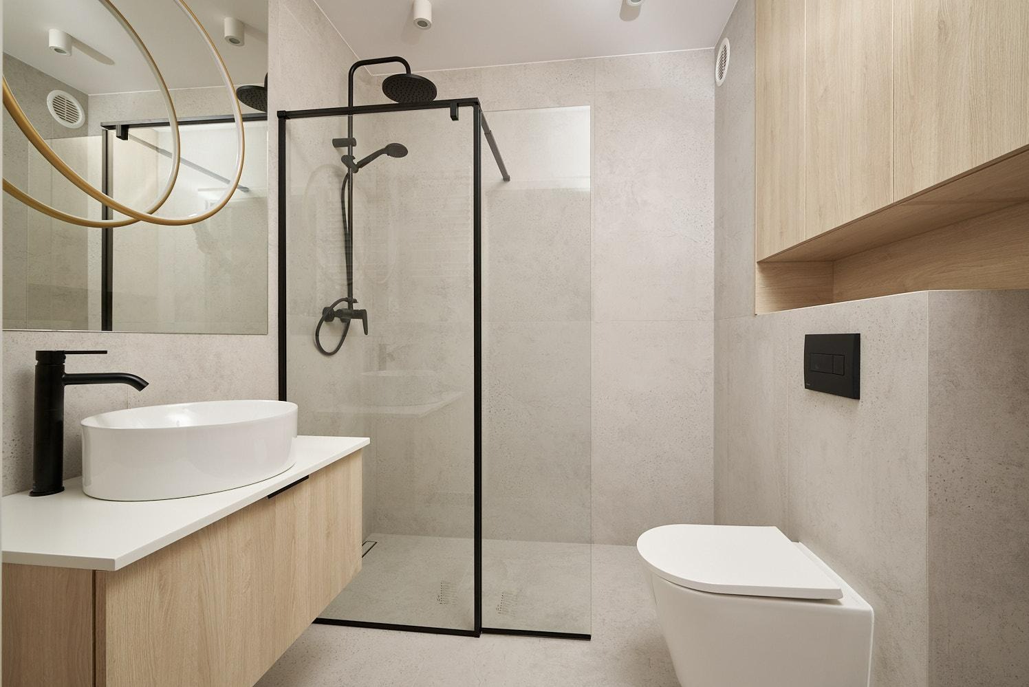 Nowoczesna łazienka z kabiną prysznicową typu walk-in z czarnymi profilami, umywalką nablatową na szafce z drewnopodobnym fornirem oraz wiszącą toaletą z czarnym przyciskiem spłukującym.