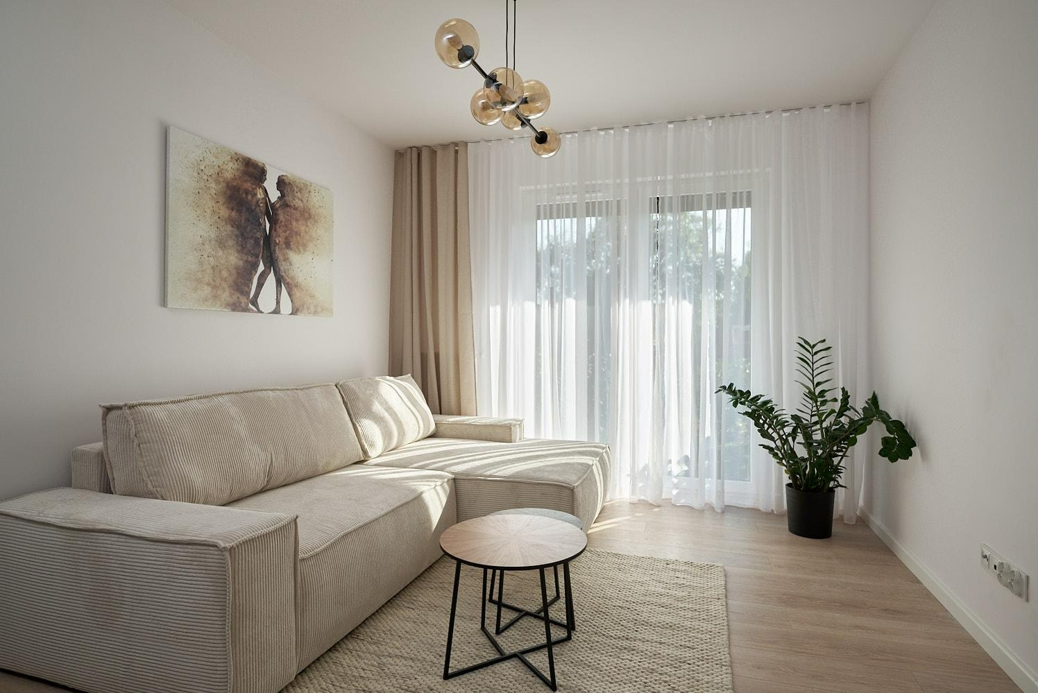 Jasny salon z narożną sofą z szerokim sztruksem, okrągłym stolikiem kawowym na metalowej podstawie, abstrakcyjnym obrazem na ścianie i dekoracyjną lampą wiszącą z kulistymi kloszami