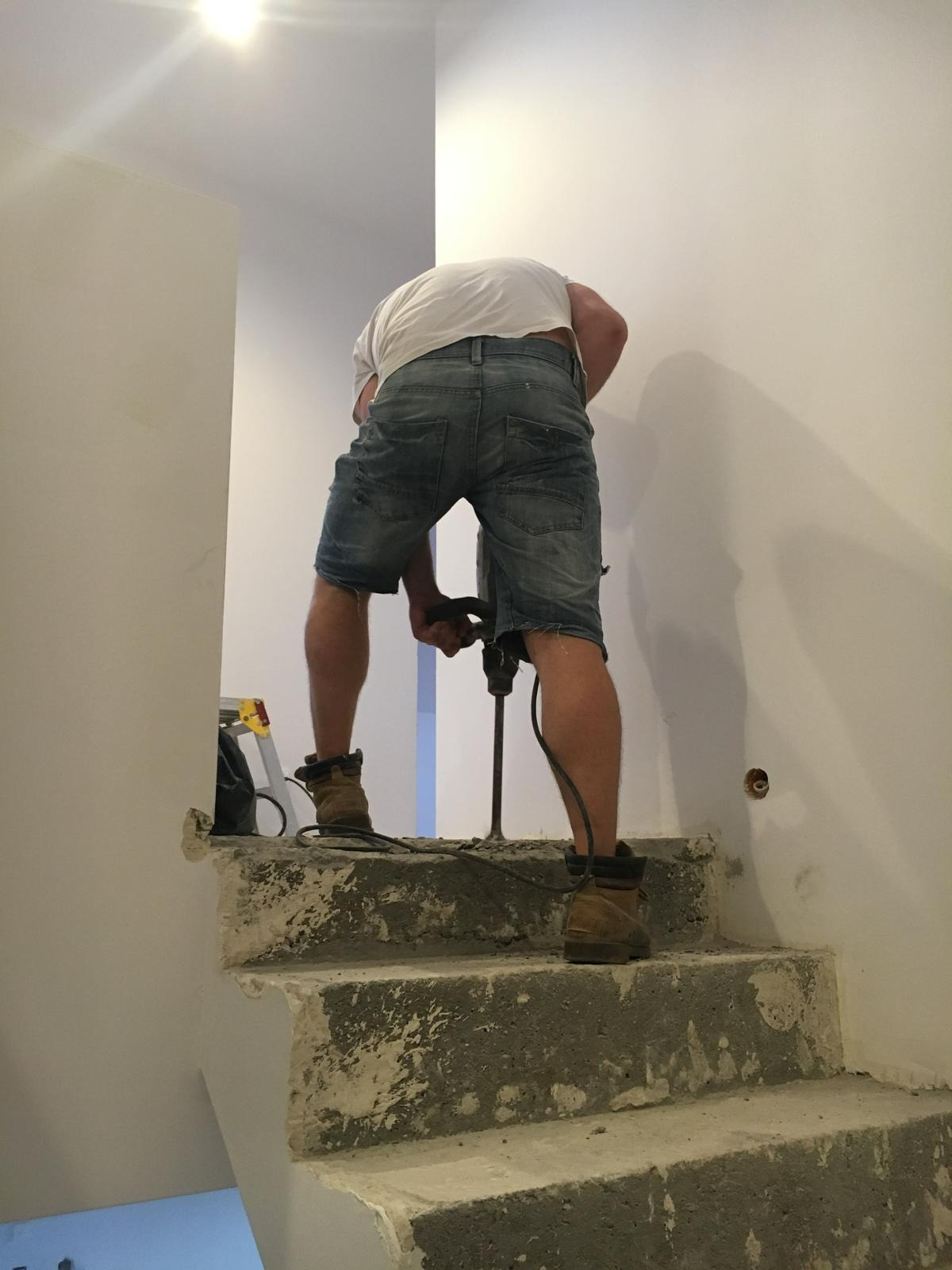 Mężczyzna w dżinsowych szortach wierci w ścianie na schodach, widoczny stary beton i przygotowania do remontu.