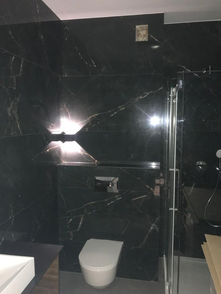 Nowoczesna łazienka z czarnymi płytkami imitującymi marmur, podświetlana ściana, wisząca toaleta i szklana kabina prysznicowa.