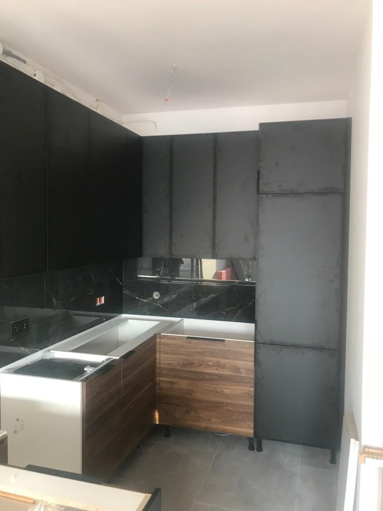 Częściowo zamontowana kuchnia z czarnymi szafkami wiszącymi i stojącymi oraz drewnopodobnymi frontami szafek dolnych, marmurowy backsplash, na podłodze szare płytki, widoczne elementy instalacji...