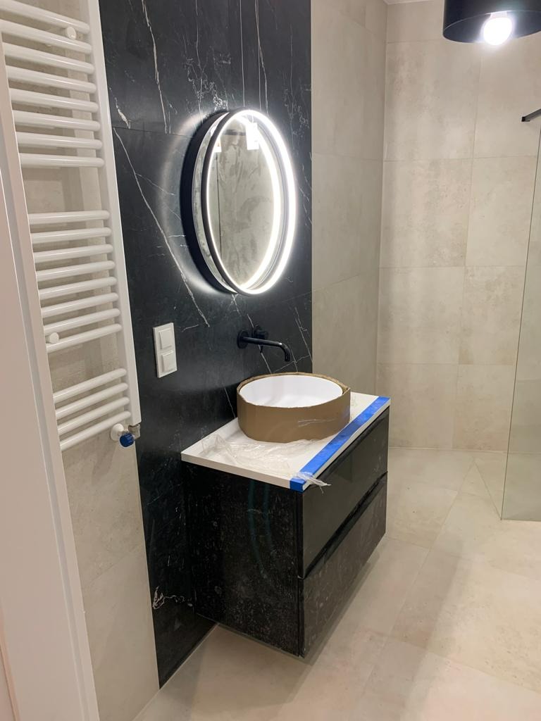 Nowoczesna łazienka w trakcie wykańczania: czarna ściana z marmurowym wzorem, okrągłe lustro z podświetleniem LED, szafka pod umywalkę z czarnego kamienia, biały grzejnik drabinkowy, umywalka...