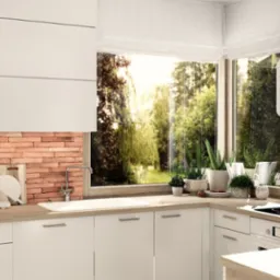 Jasna kuchnia z białymi szafkami, ceglaną ścianą nad blatem, widokiem na zieleń za oknem i robotem kuchennym Thermomix na blacie.