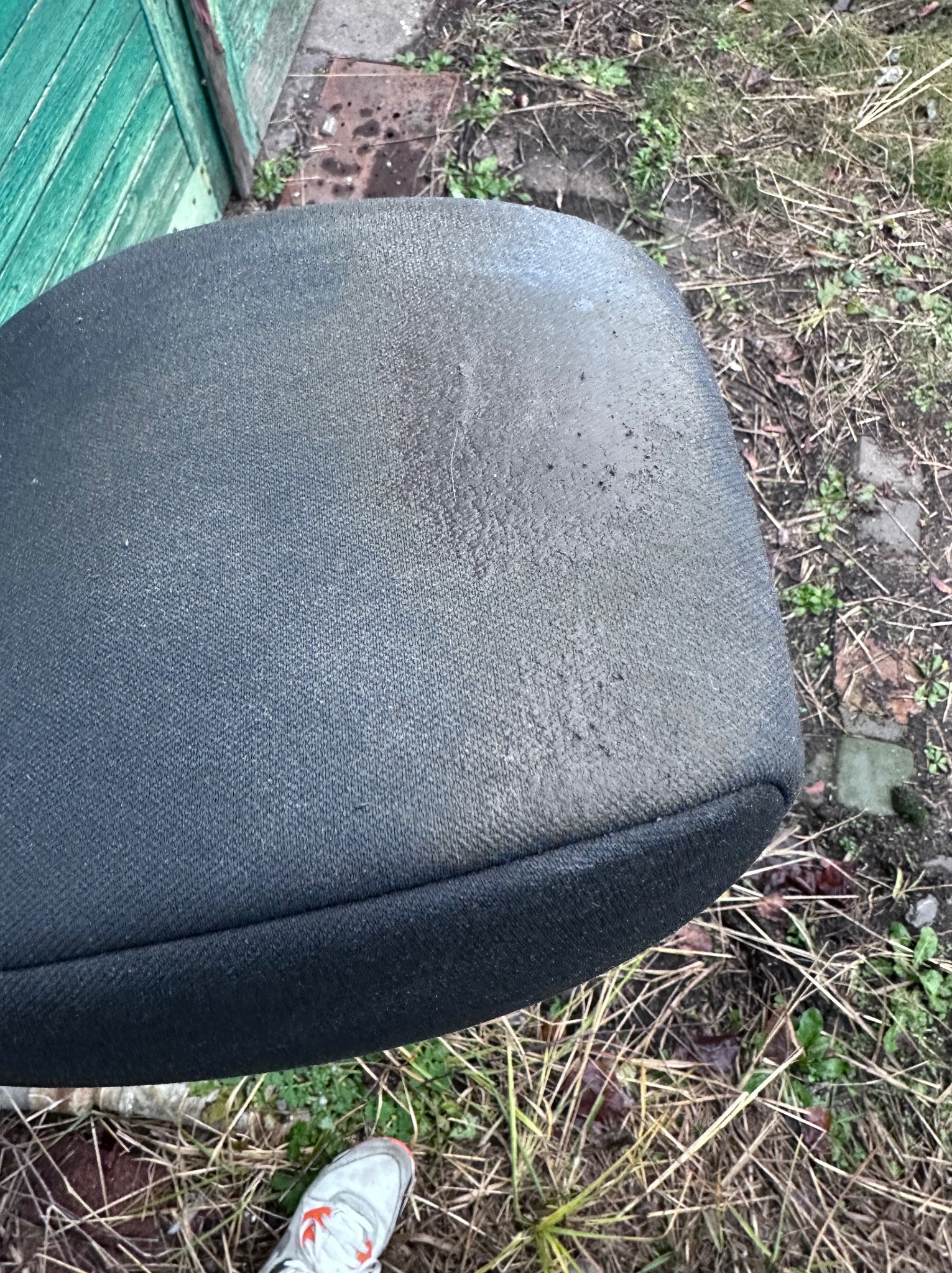 Brudne siedzisko krzesła biurowego z szarej tkaniny, widoczne plamy i zabrudzenia, tło z zielonymi deskami i ziemią z suchą trawą, u dołu fragment buta sportowego.