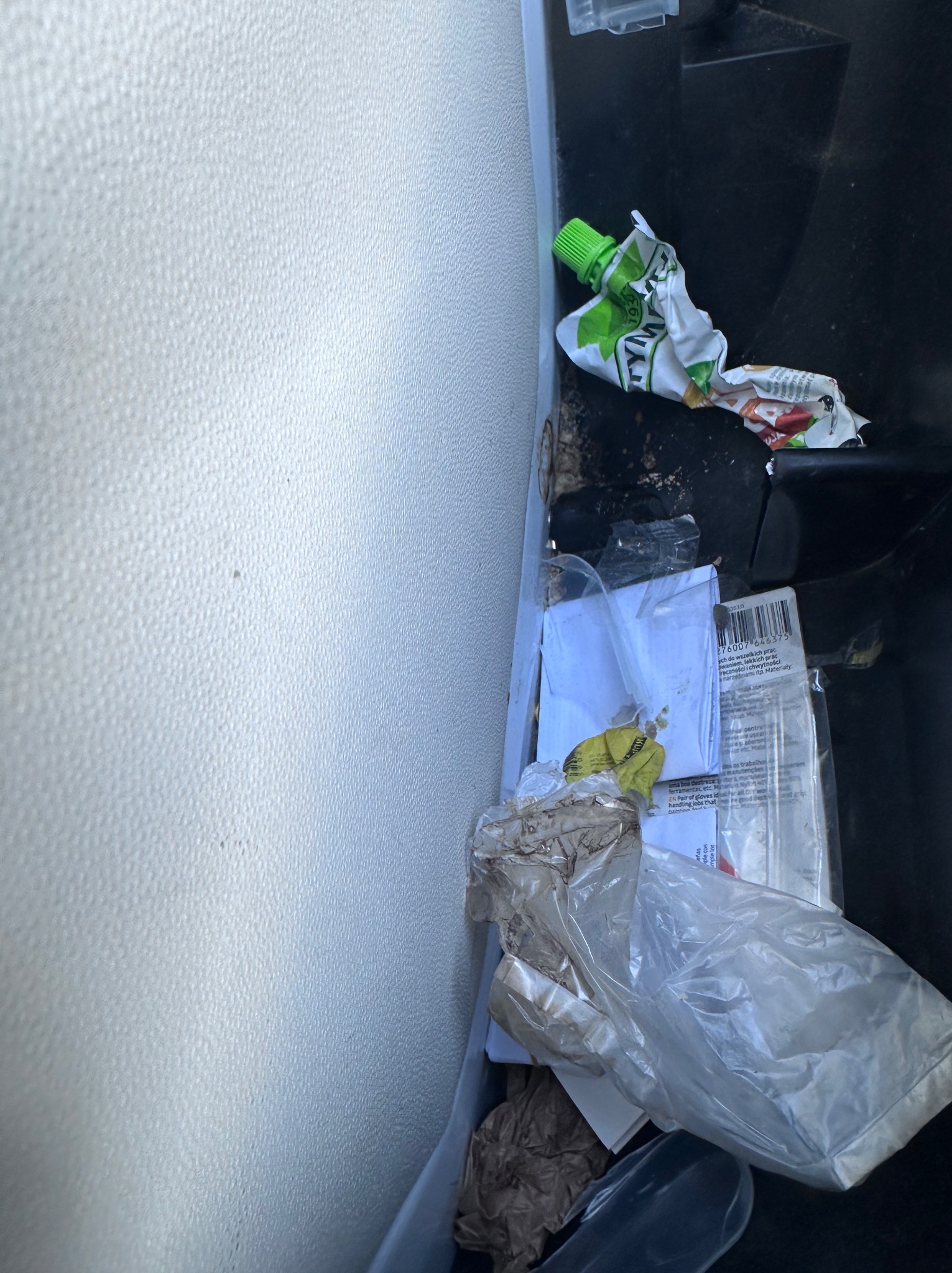 Niezorganizowany schowek samochodowy wypełniony śmieciami, w tym zużyte opakowania po żywności, plastikowe torby i dokumenty, ukazujący potrzebę sprzątania wnętrza auta.