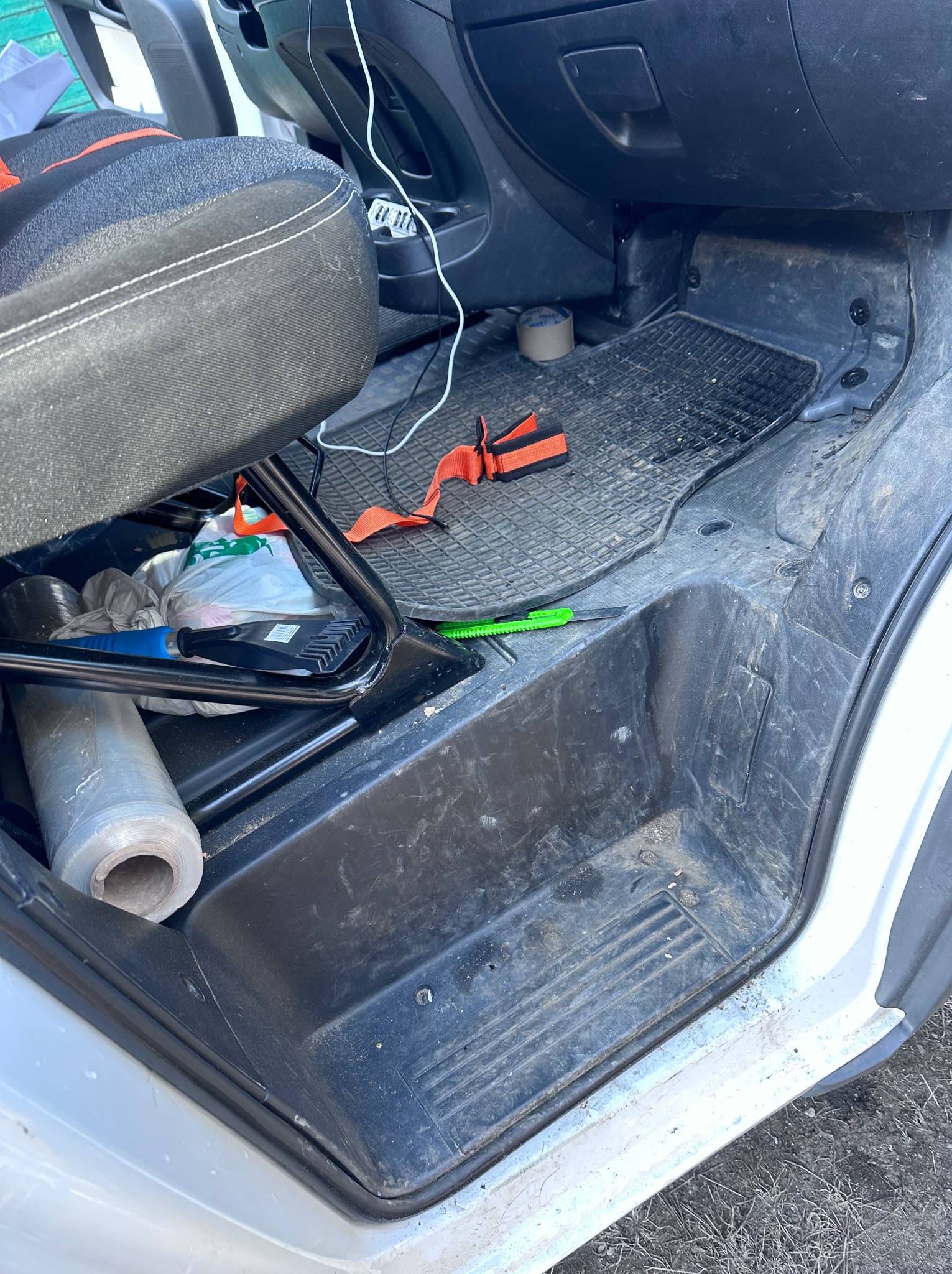 Wnętrze brudnego busa z widocznym siedziskiem, podłogą z gumową wycieraczką i narzędziami. W tle deska rozdzielcza i kabel. Widoczne ślady użytkowania.