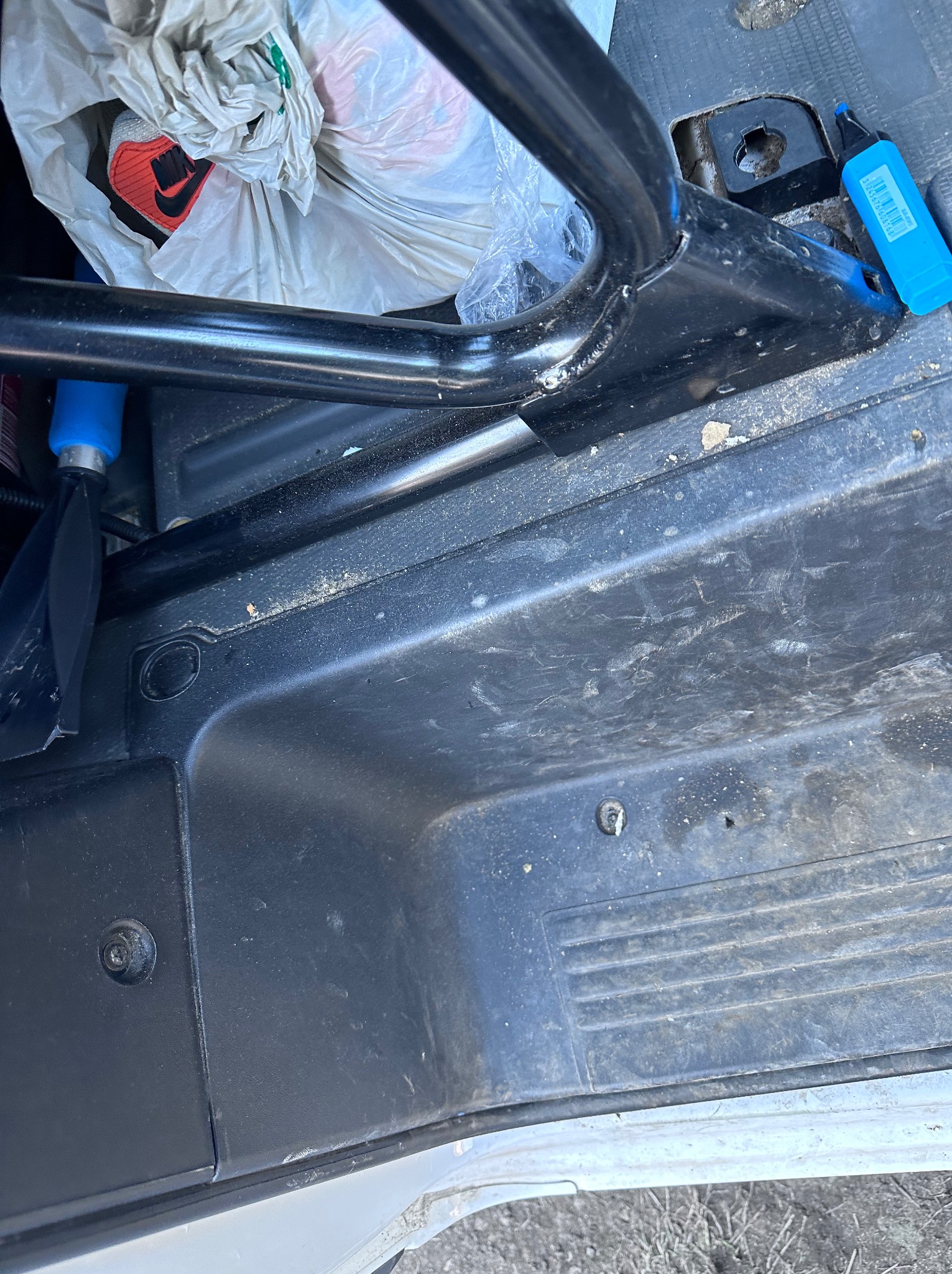 Wnętrze brudnego busa transportowego z czarną metalową konstrukcją, plastikową podłogą pokrytą piaskiem i śmieciami oraz torbą z obuwiem w tle.