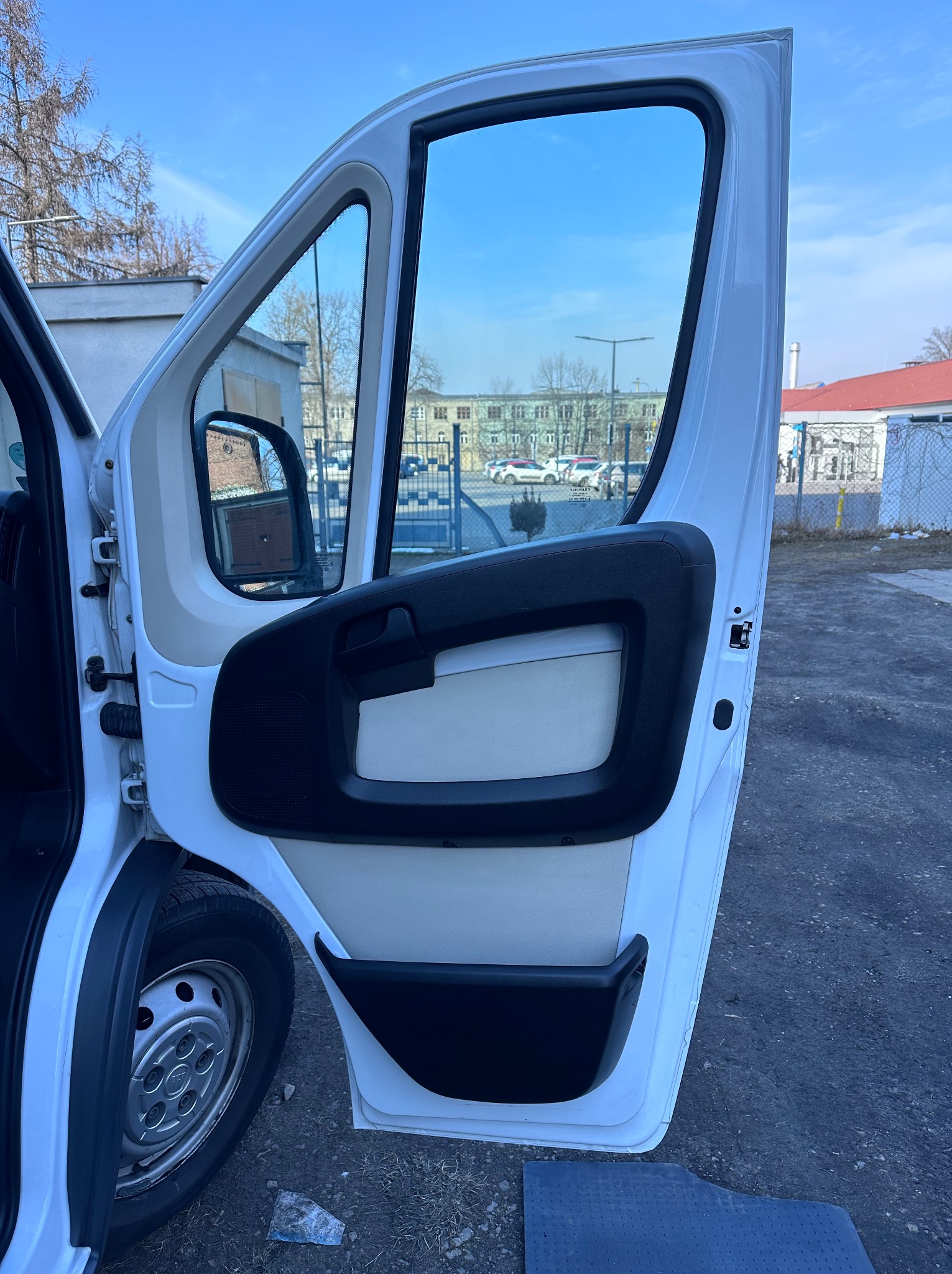 Otwarta boczna drzwi białego busa dostawczego z widocznym wnętrzem, oponą i fragmentem otoczenia miejskiego w tle.