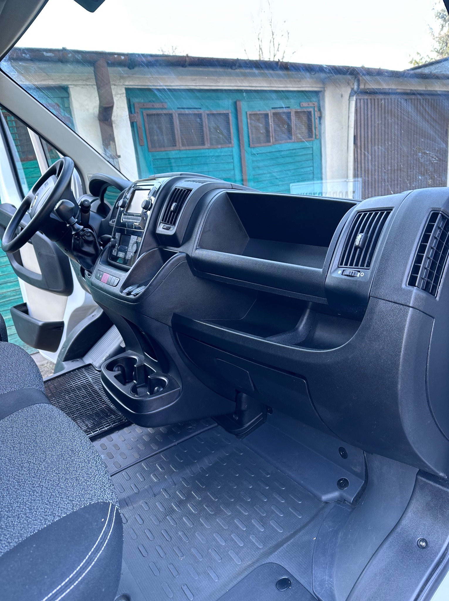 Wnętrze busa: czarna deska rozdzielcza, kratki wentylacyjne, schowki, uchwyty na napoje, gumowa wycieraczka i fragment fotela kierowcy. W tle budynek z niebieskimi drzwiami.