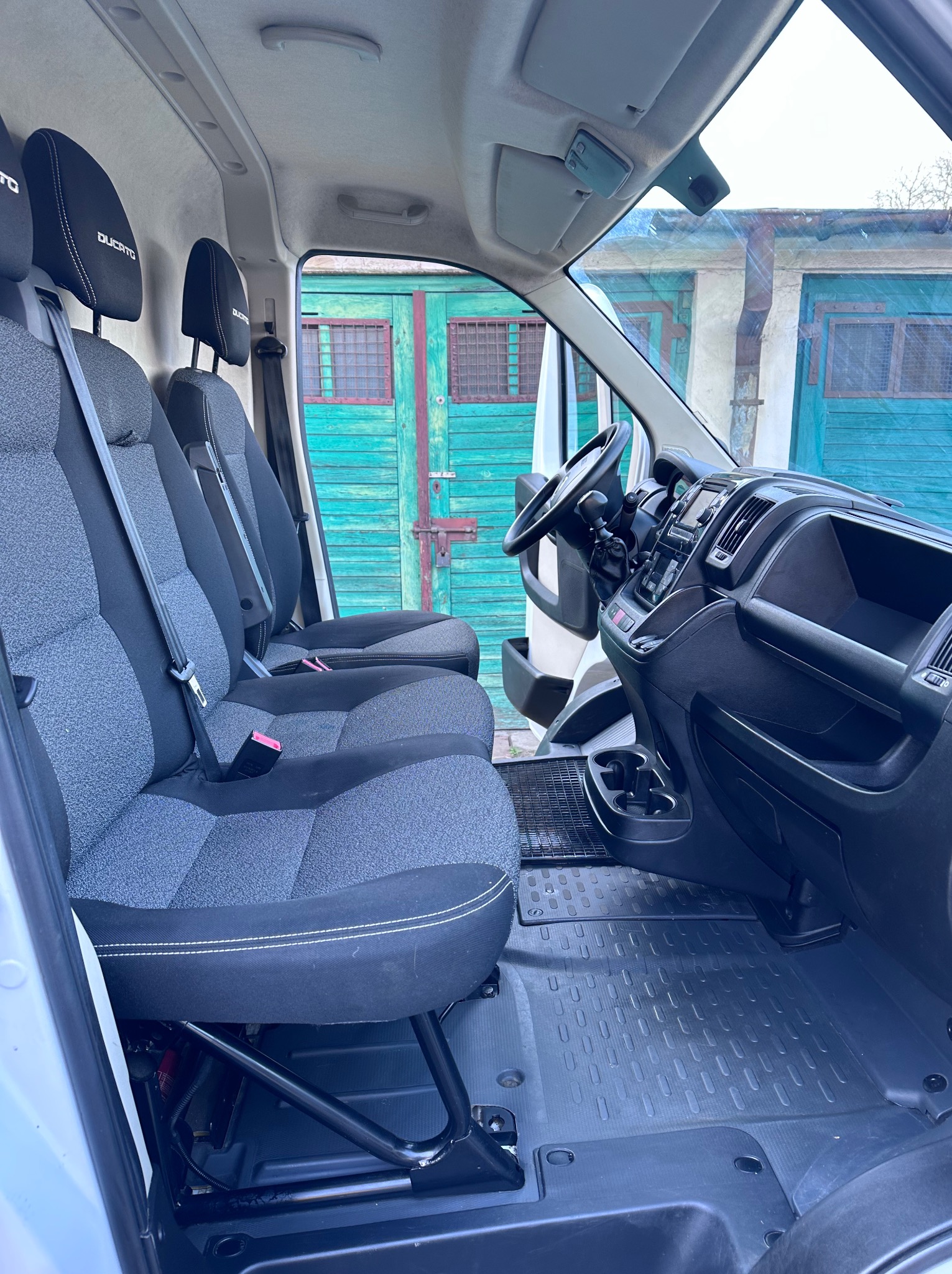 Wnętrze busa z trzema fotelami z szarej tapicerki i czarnymi zagłówkami, widok na deskę rozdzielczą i zielone drzwi garażowe w tle.