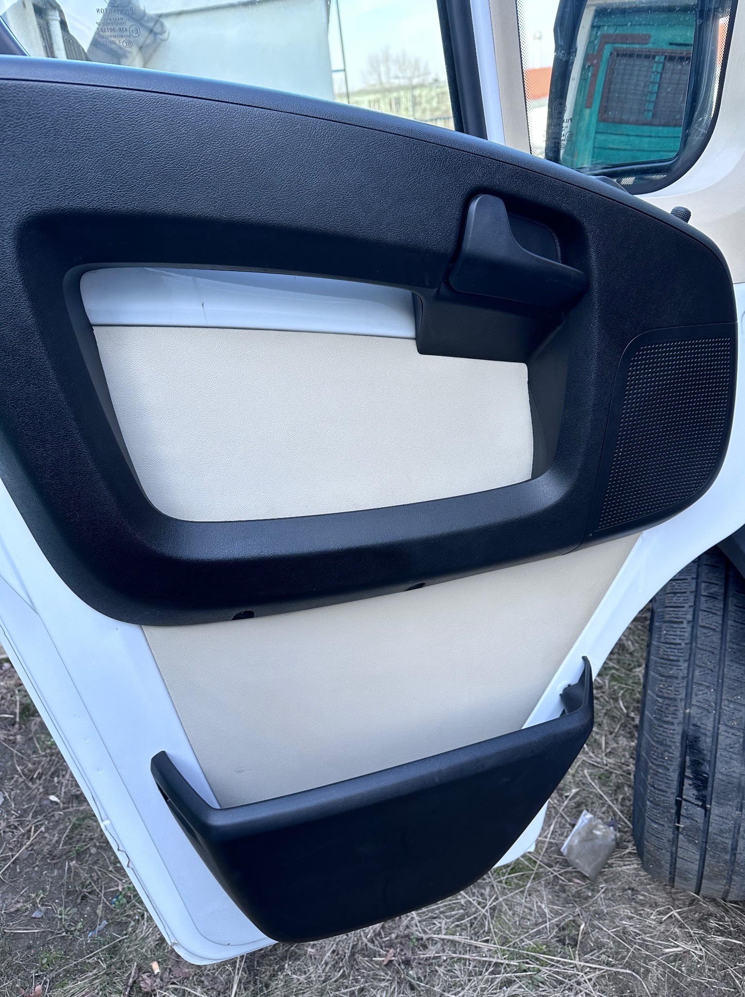 Wnętrze drzwi pojazdu z czarną plastikową ramą i beżową tapicerką. Widoczne fragment opony i szyby.