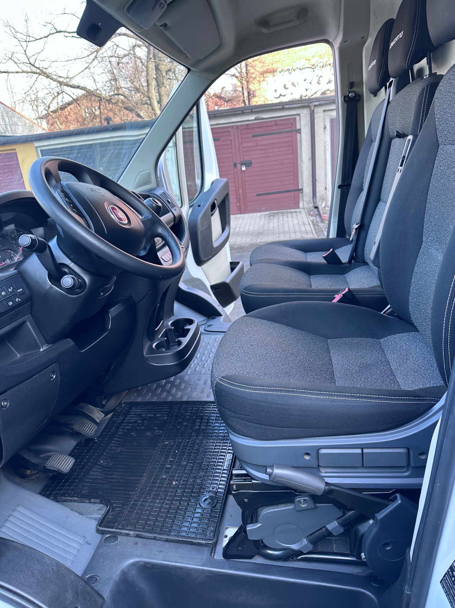 Wnętrze białego busa marki Fiat Ducato z widocznymi fotelami kierowcy i pasażerów, czarnymi dywanikami gumowymi oraz elementami deski rozdzielczej.
