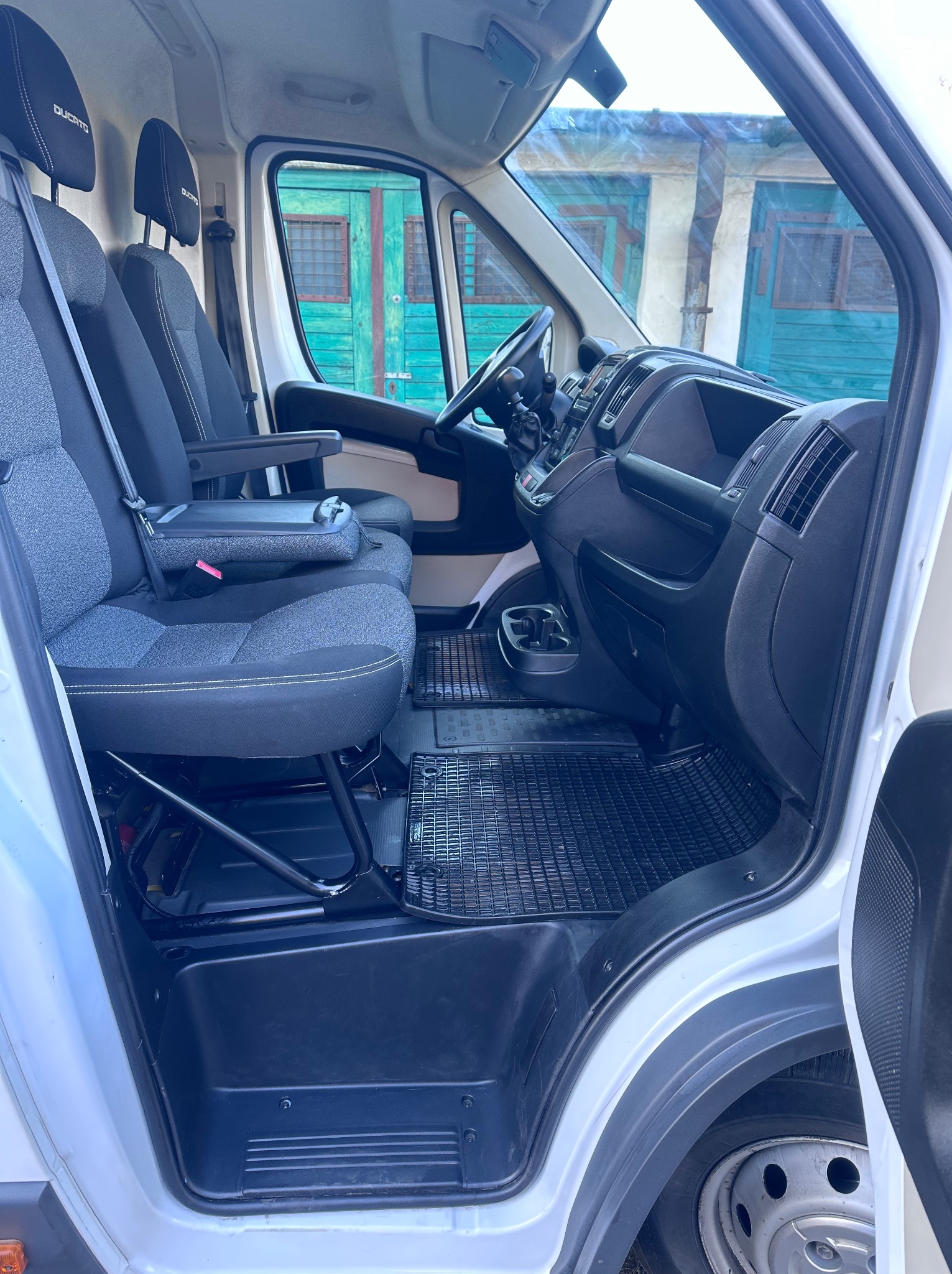 Wnętrze busa marki Ducato z szarymi fotelami i czarnymi elementami deski rozdzielczej, widok z boku przez otwarte drzwi, w tle budynek z turkusowymi drzwiami.
