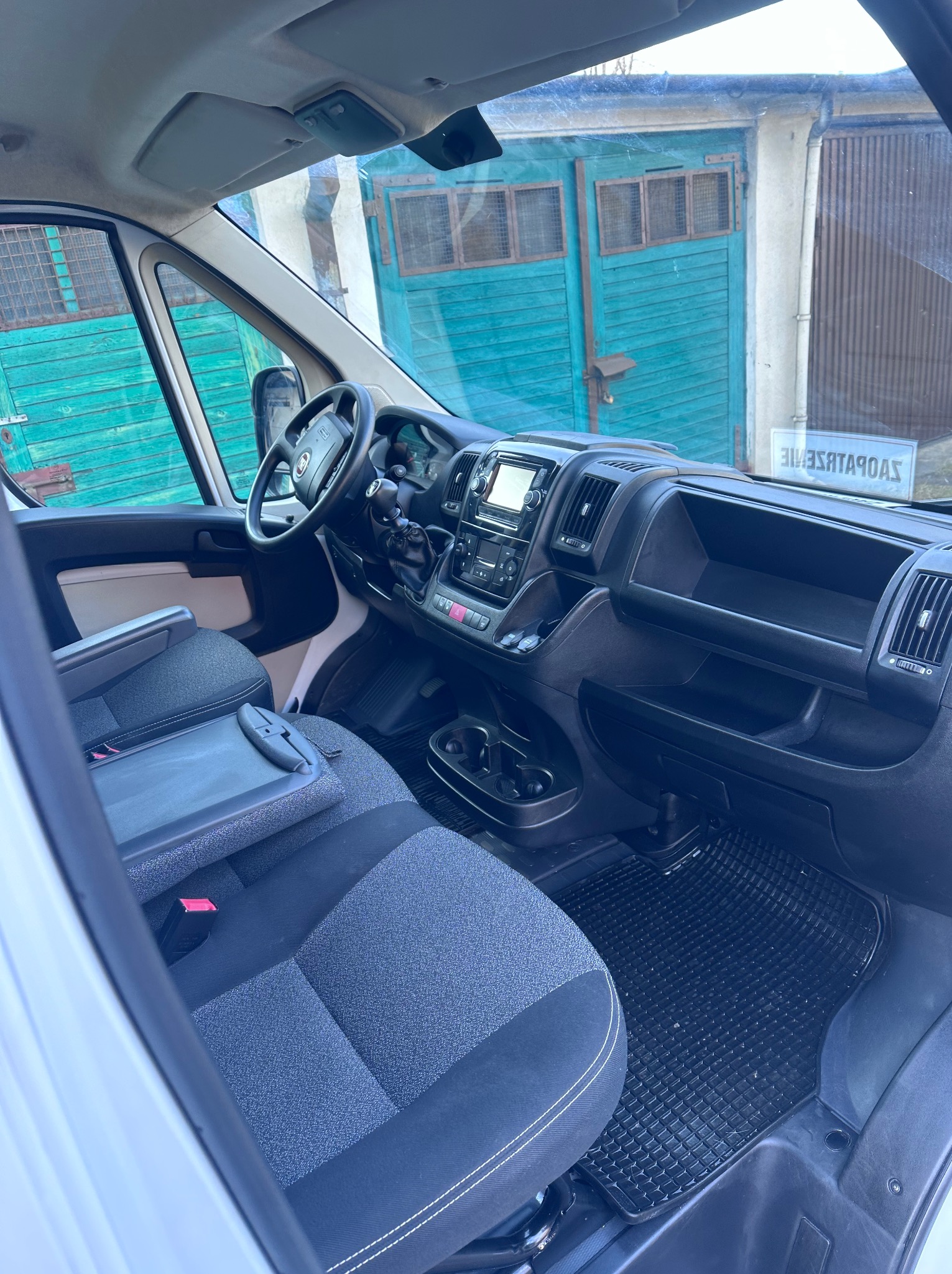 Wnętrze busa z szarą tapicerką i rozkładanym stolikiem, widok z perspektywy pasażera, na tle garażu z turkusowymi drzwiami.