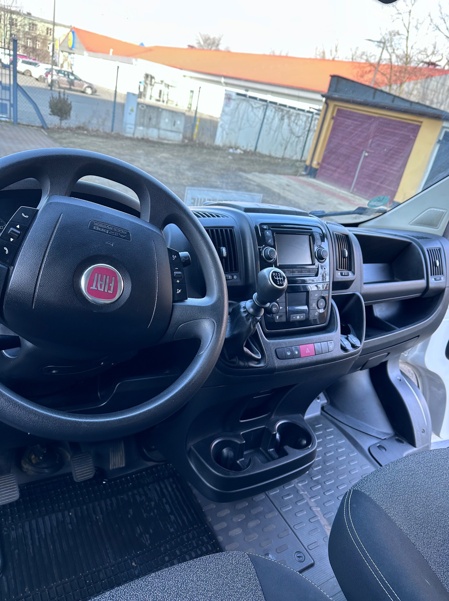 Wnętrze kabiny busa marki Fiat z widoczną deską rozdzielczą, kierownicą z logo, dźwignią zmiany biegów i siedzeniem kierowcy. Tło stanowią budynki.