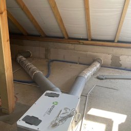 Mateusz Góral - Centrala wentylacyjna z rekuperacją Regnet zamontowana na poddaszu, widoczne przewody wentylacyjne, drewniana konstrukcja dachu i surowe ściany z betonu komórkowego.