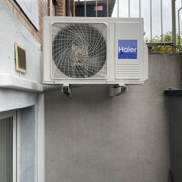 Mateusz Góral - Zewnętrzna jednostka klimatyzacji Haier zamontowana na szarej ścianie budynku, widoczne metalowe wsporniki i peszel odprowadzający skropliny, na górze metalowa balustrada.