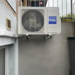 Zewnętrzna jednostka klimatyzacji Haier zamontowana na szarej ścianie budynku, widoczne metalowe wsporniki i peszel odprowadzający skropliny, na górze metalowa balustrada.