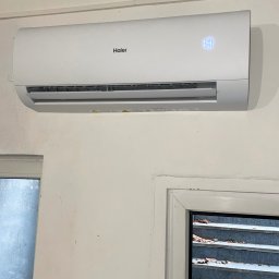 Mateusz Góral - Biały klimatyzator Haier zamontowany na ścianie z widocznym wyświetlaczem temperatury 19 stopni Celsjusza, fragment okna z widokiem na schody.