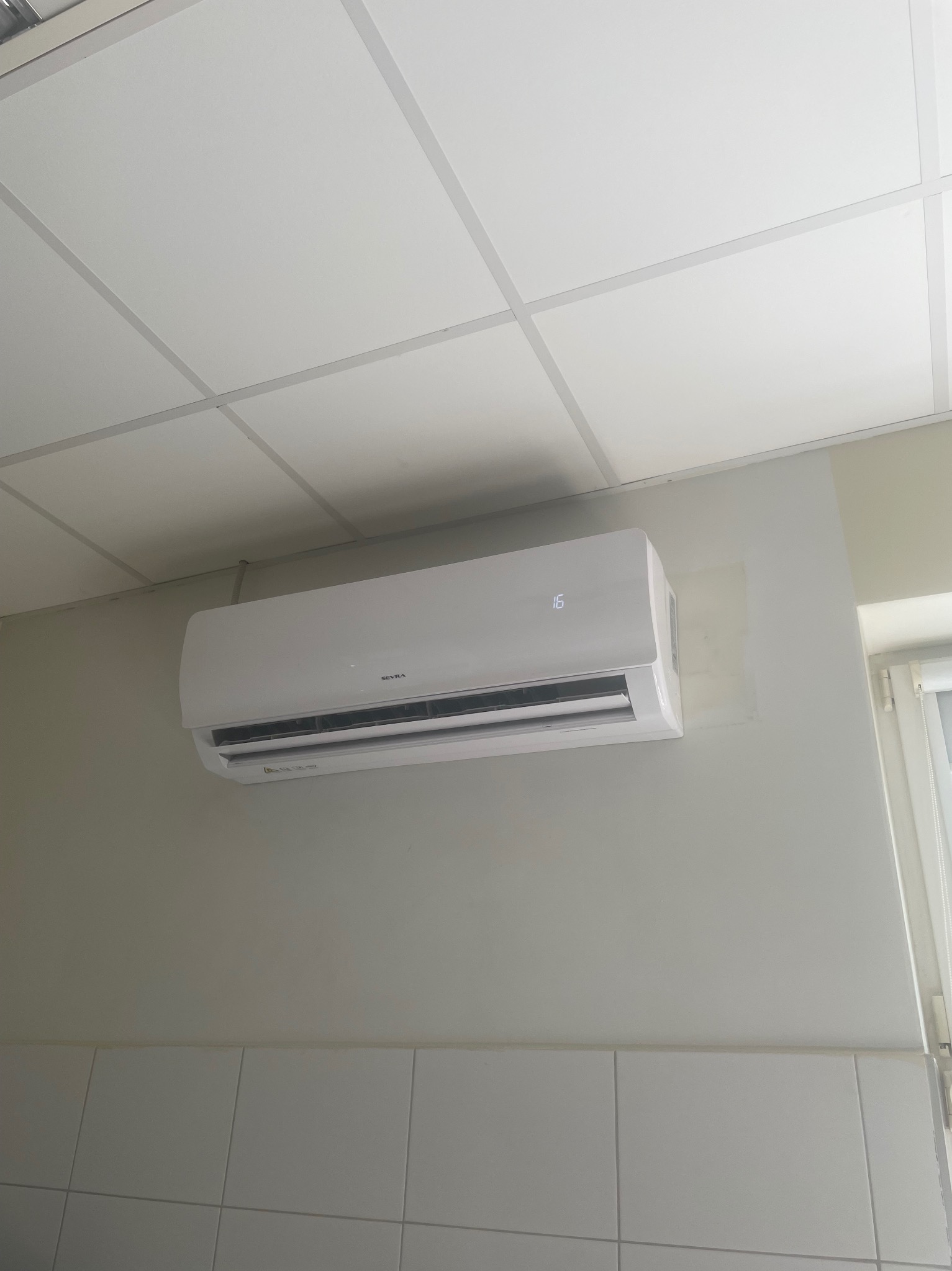 Biały klimatyzator marki Sevra zamontowany na ścianie z kafelkami, widoczny wyświetlacz z temperaturą 16 stopni, sufit kasetonowy w tle.