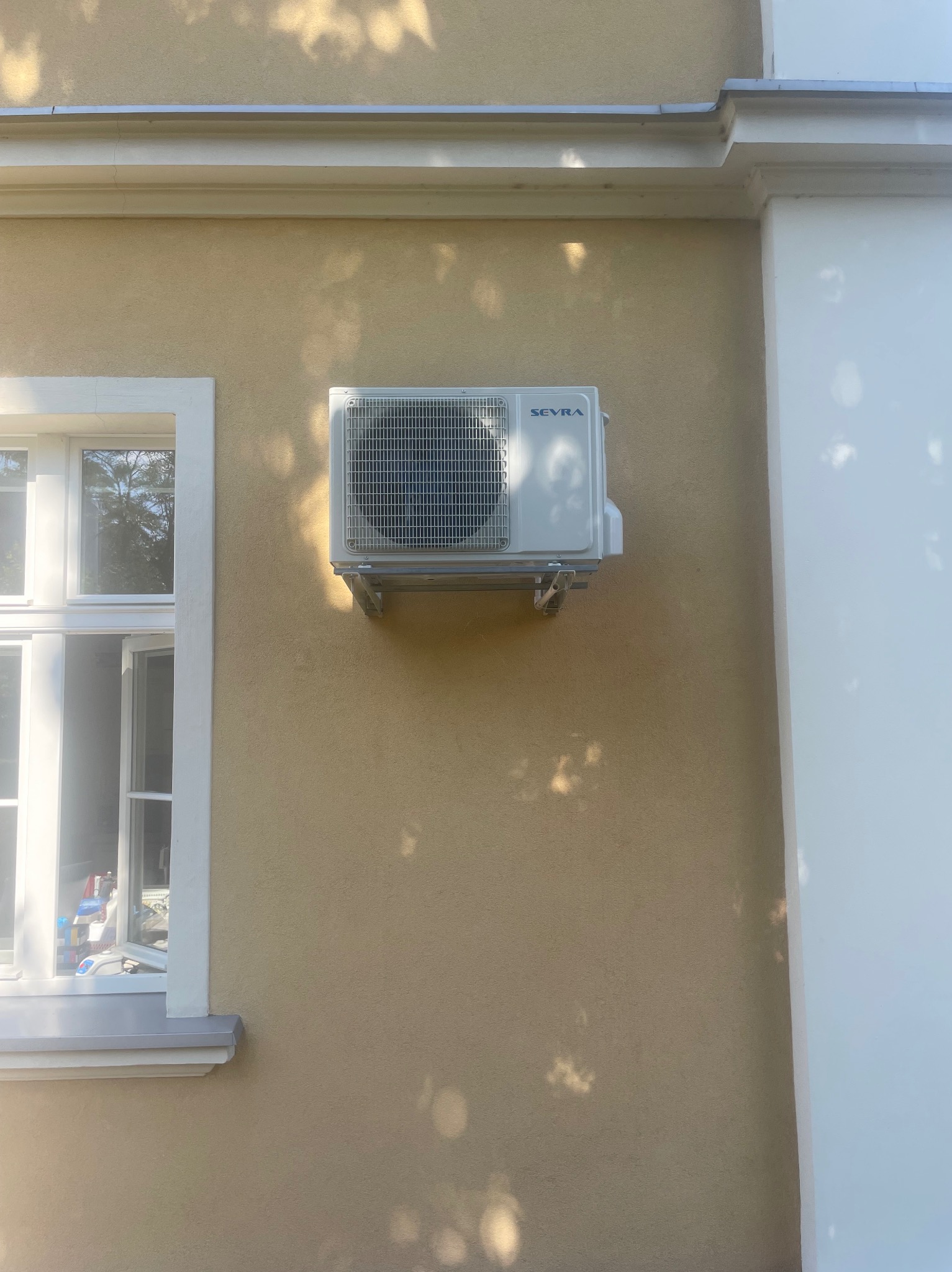 Biały klimatyzator marki Seyra zamontowany na beżowej ścianie budynku, widoczne cienie drzew i okno z białymi ramami.