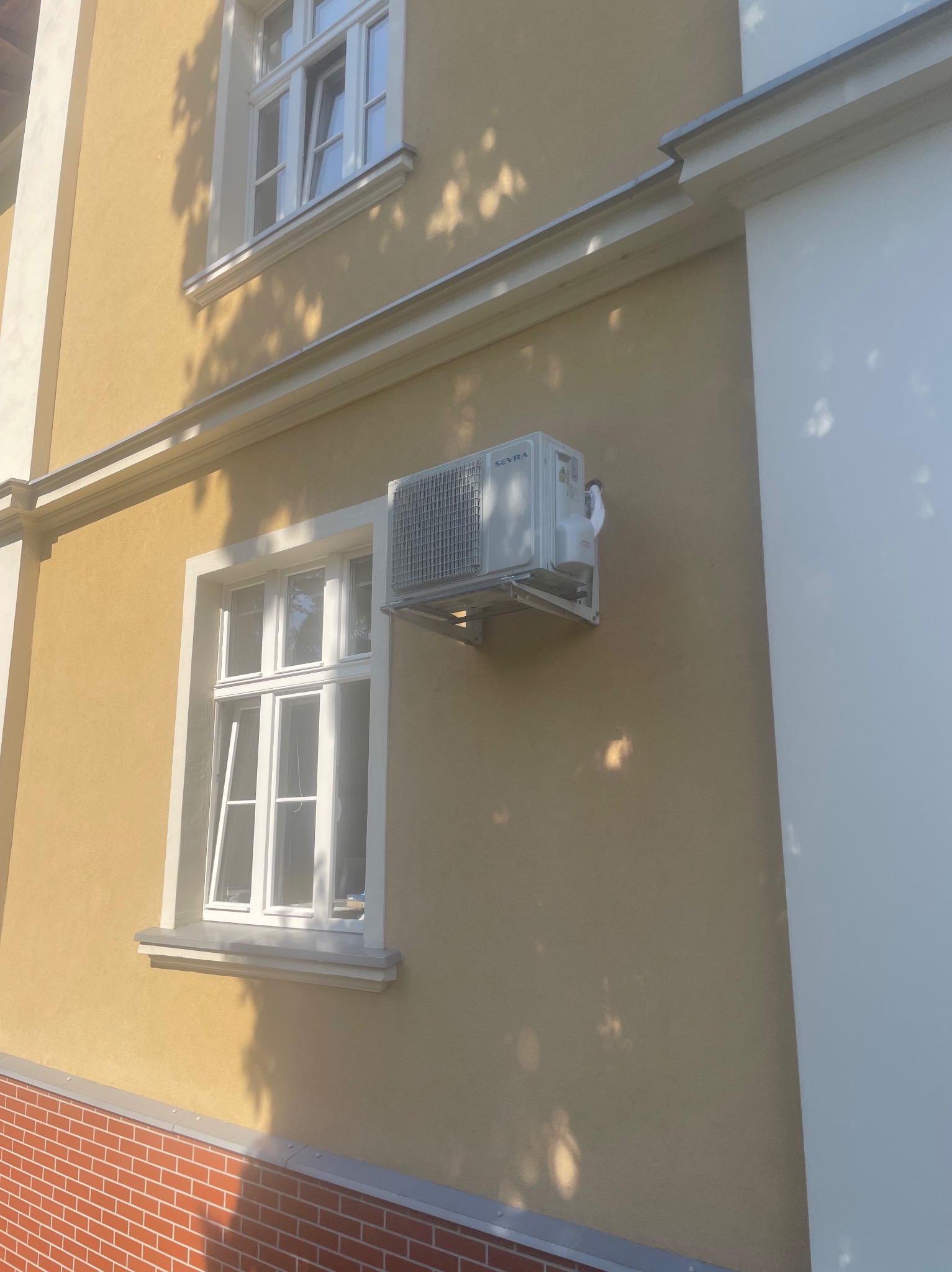 Zewnętrzna jednostka klimatyzacji zamontowana na żółtej elewacji budynku obok okna z białymi ramami.