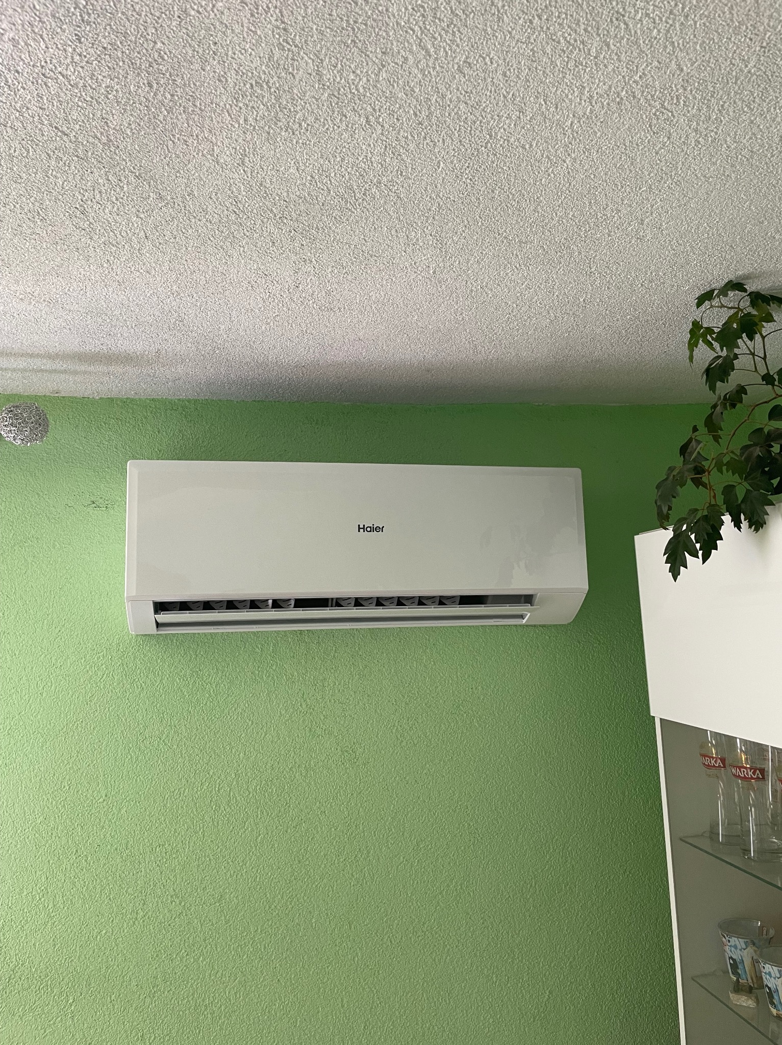Biała jednostka klimatyzacyjna Haier zamontowana na zielonej ścianie w pomieszczeniu z widoczną rośliną doniczkową i fragmentem przeszklonej witryny.