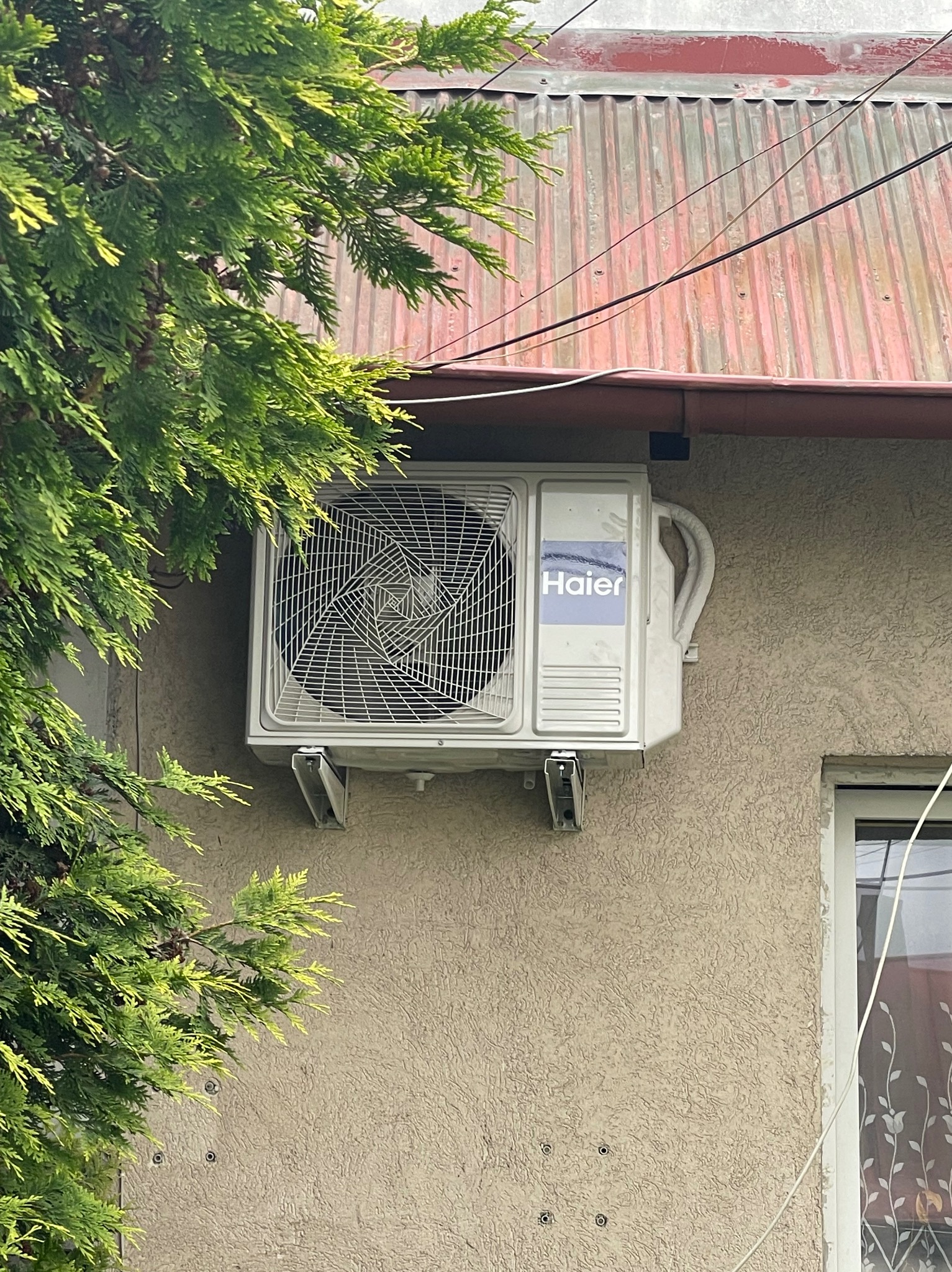 Biała jednostka zewnętrzna klimatyzatora Haier zamontowana na elewacji domu o beżowej fakturze, widoczna spod gałęzi drzewa, z fragmentem okna po prawej stronie.