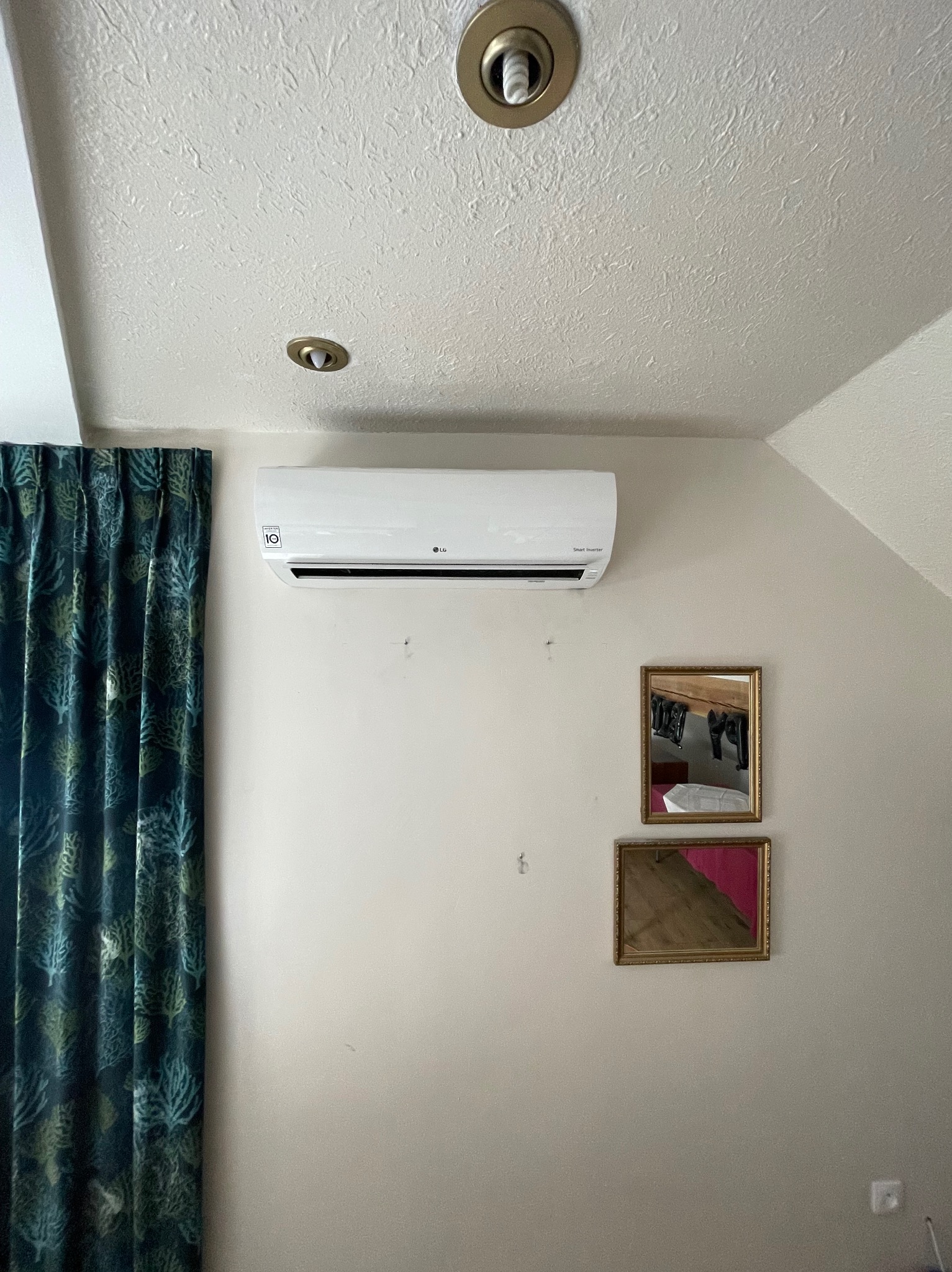 Biała jednostka klimatyzacji LG zamontowana na ścianie w pokoju z zasłonami w odcieniach zieleni i dwoma lustrami w złotych ramach.