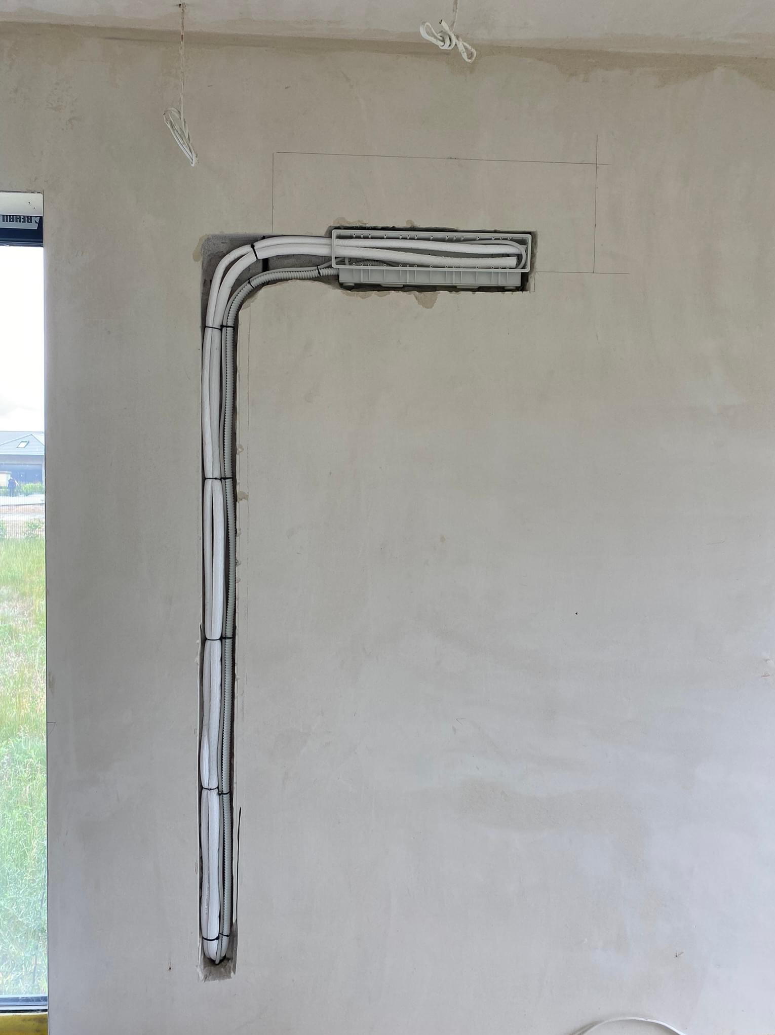 Instalacja przewodów w ścianie podczas remontu, widoczne kable w białych peszlach ułożone w pionowym kanale i doprowadzone do puszki pod sufitem, surowy tynk na ścianie, okno w tle.