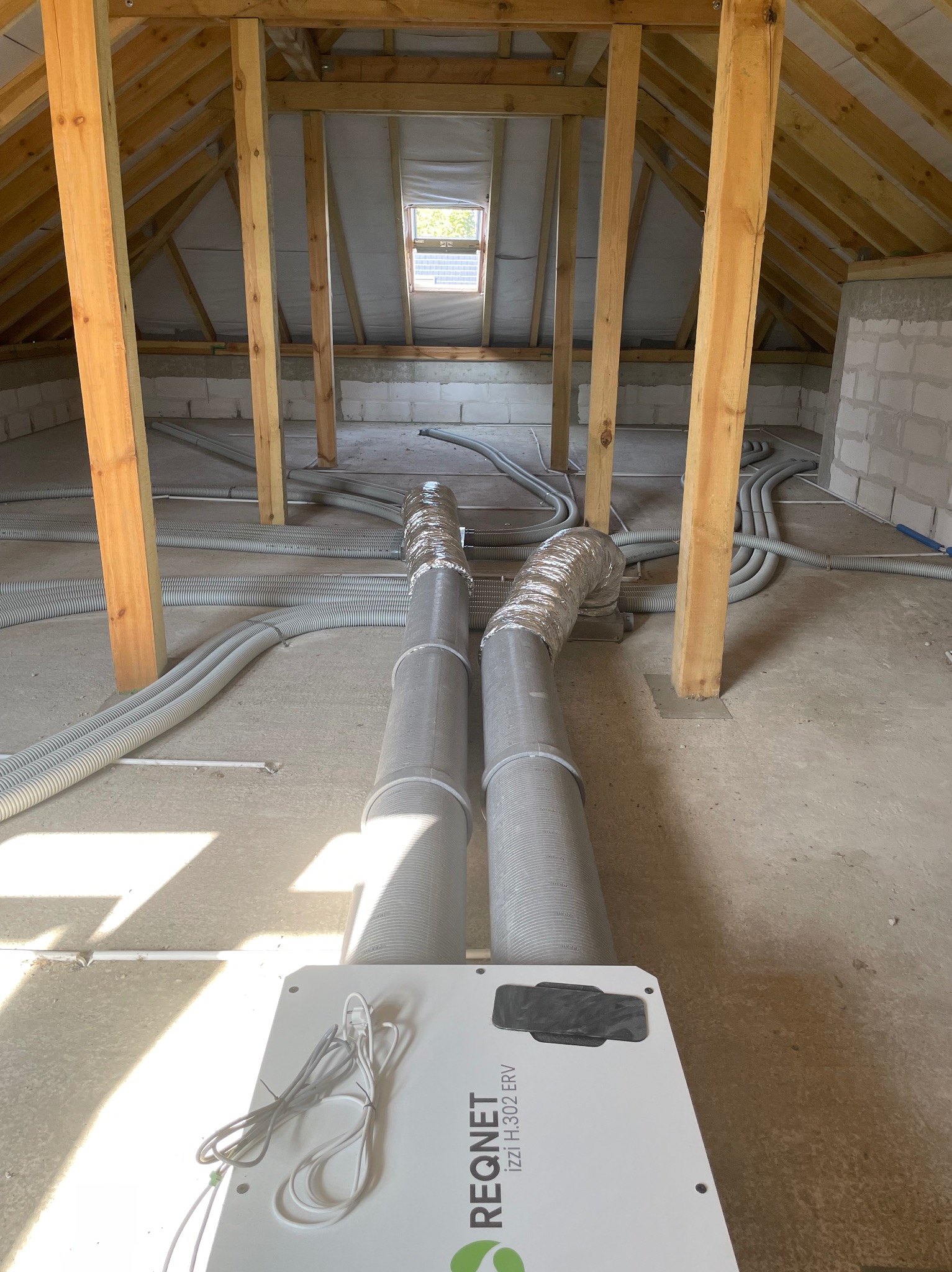 Montaż rekuperatora REQNET izzi H.302 ERV na poddaszu z widocznymi przewodami wentylacyjnymi i drewnianą konstrukcją dachu.