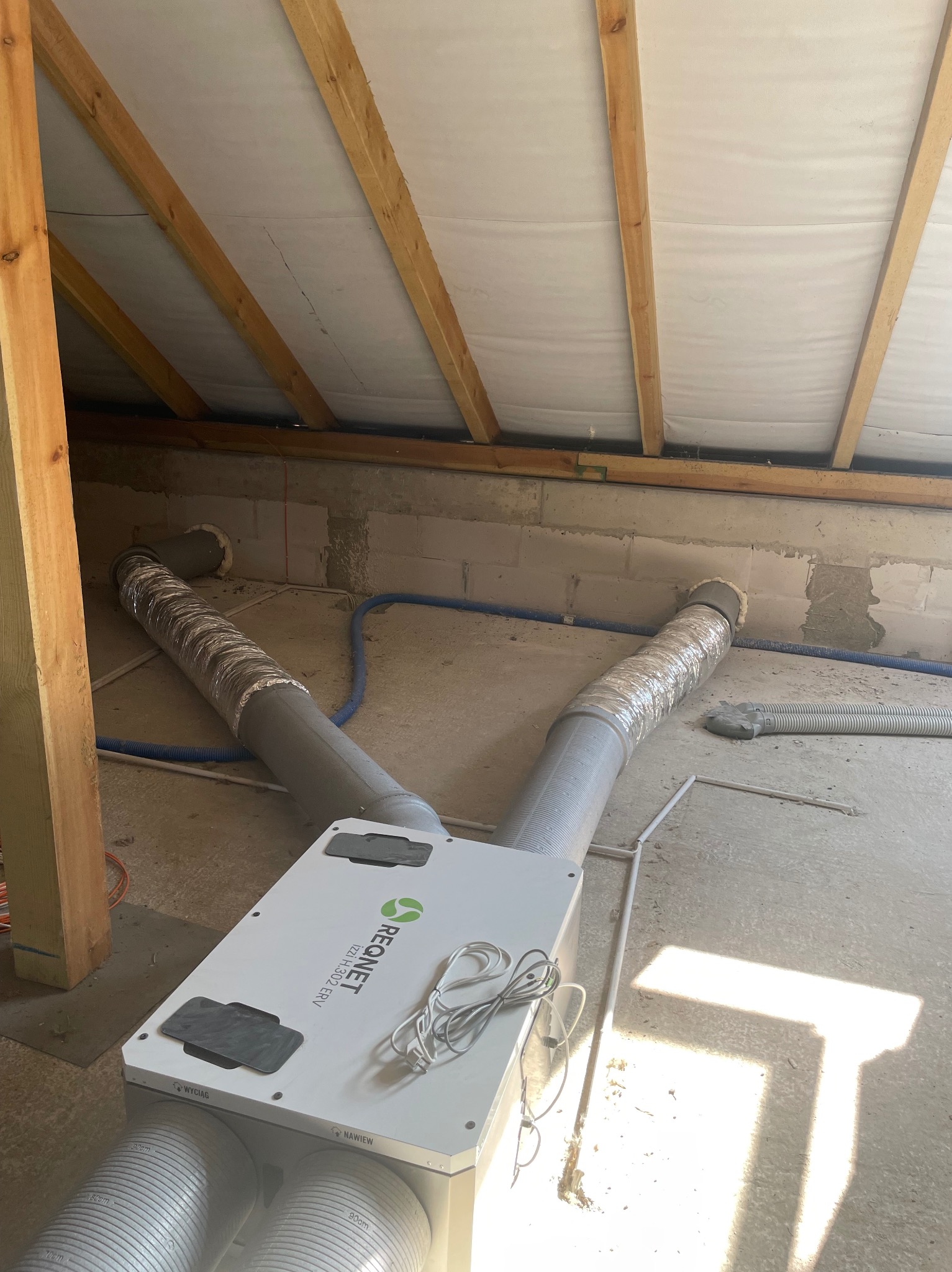 Centrala wentylacyjna z rekuperacją Regnet zamontowana na poddaszu, widoczne przewody wentylacyjne, drewniana konstrukcja dachu i surowe ściany z betonu komórkowego.