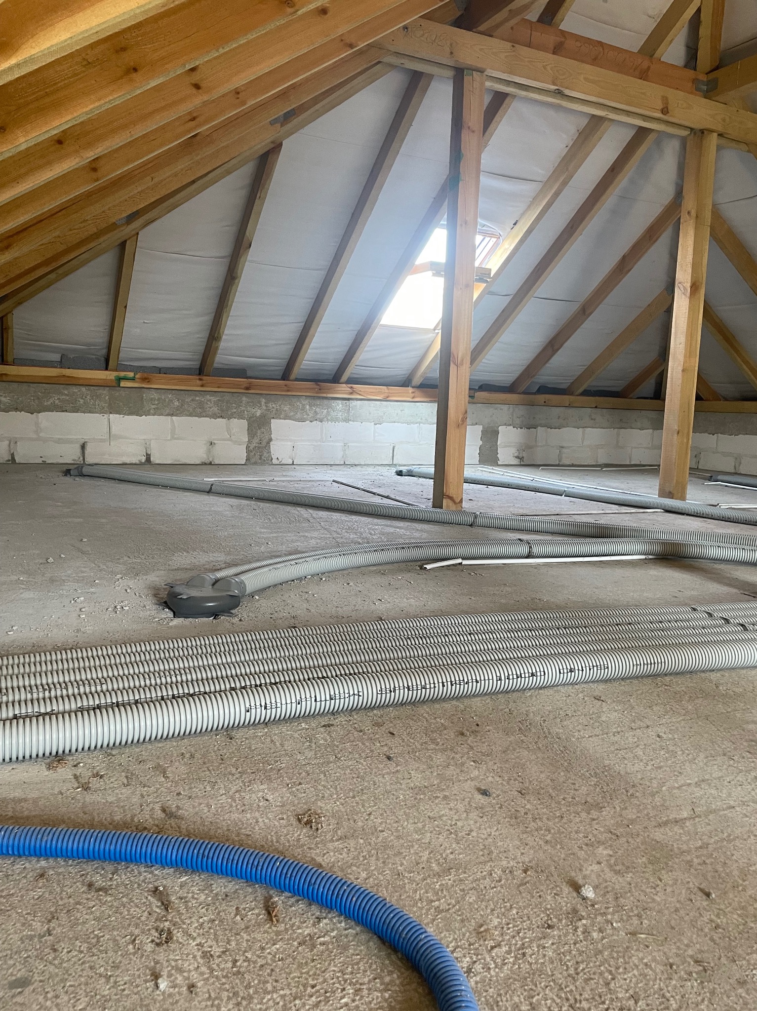 Poddasze w trakcie instalacji systemu rekuperacji: widoczne rury wentylacyjne, drewniana konstrukcja dachu i okno dachowe.