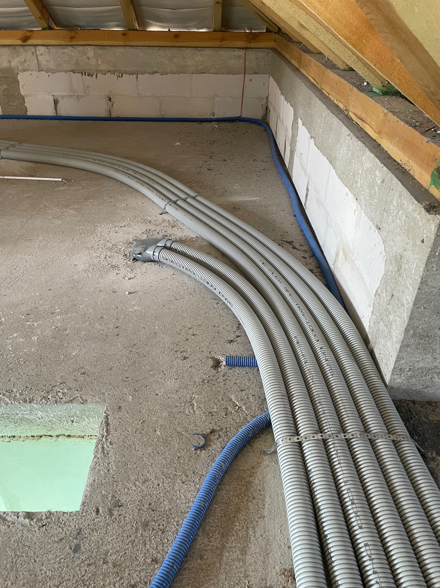 Szare rury karbowane ułożone równolegle na surowym betonie, niebieska rura wzdłuż ściany z betonu komórkowego, widoczne drewniane elementy konstrukcji dachu.