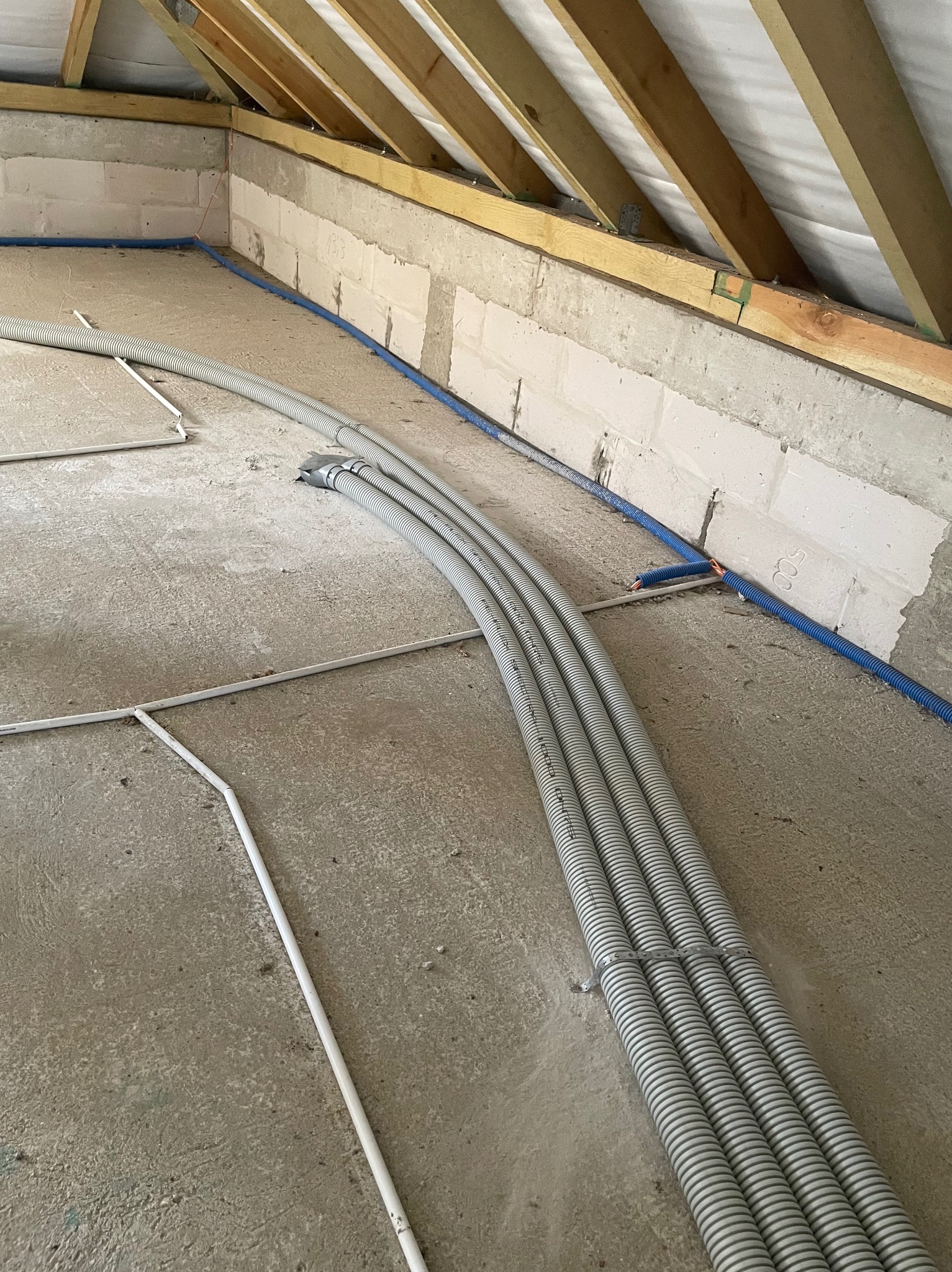 Szare, karbowane rury wentylacyjne ułożone na betonowej podłodze poddasza, przymocowane metalową obejmą, z widoczną konstrukcją dachu i fragmentem instalacji elektrycznej w niebieskiej rurce.