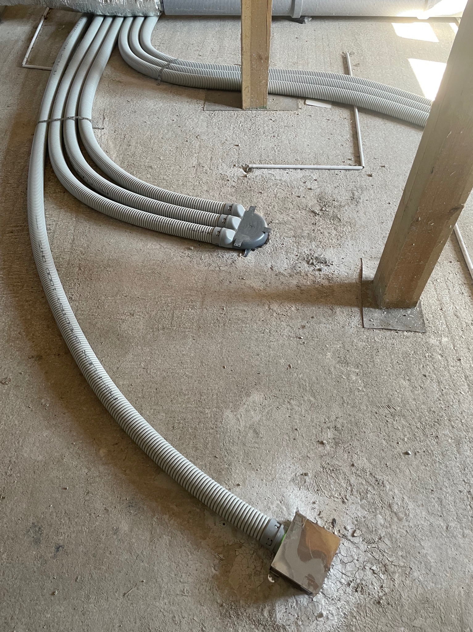 Szare, karbowane rury wentylacyjne ułożone na surowej betonowej podłodze w trakcie montażu systemu rekuperacji w budynku, widoczne drewniane podpory konstrukcyjne.