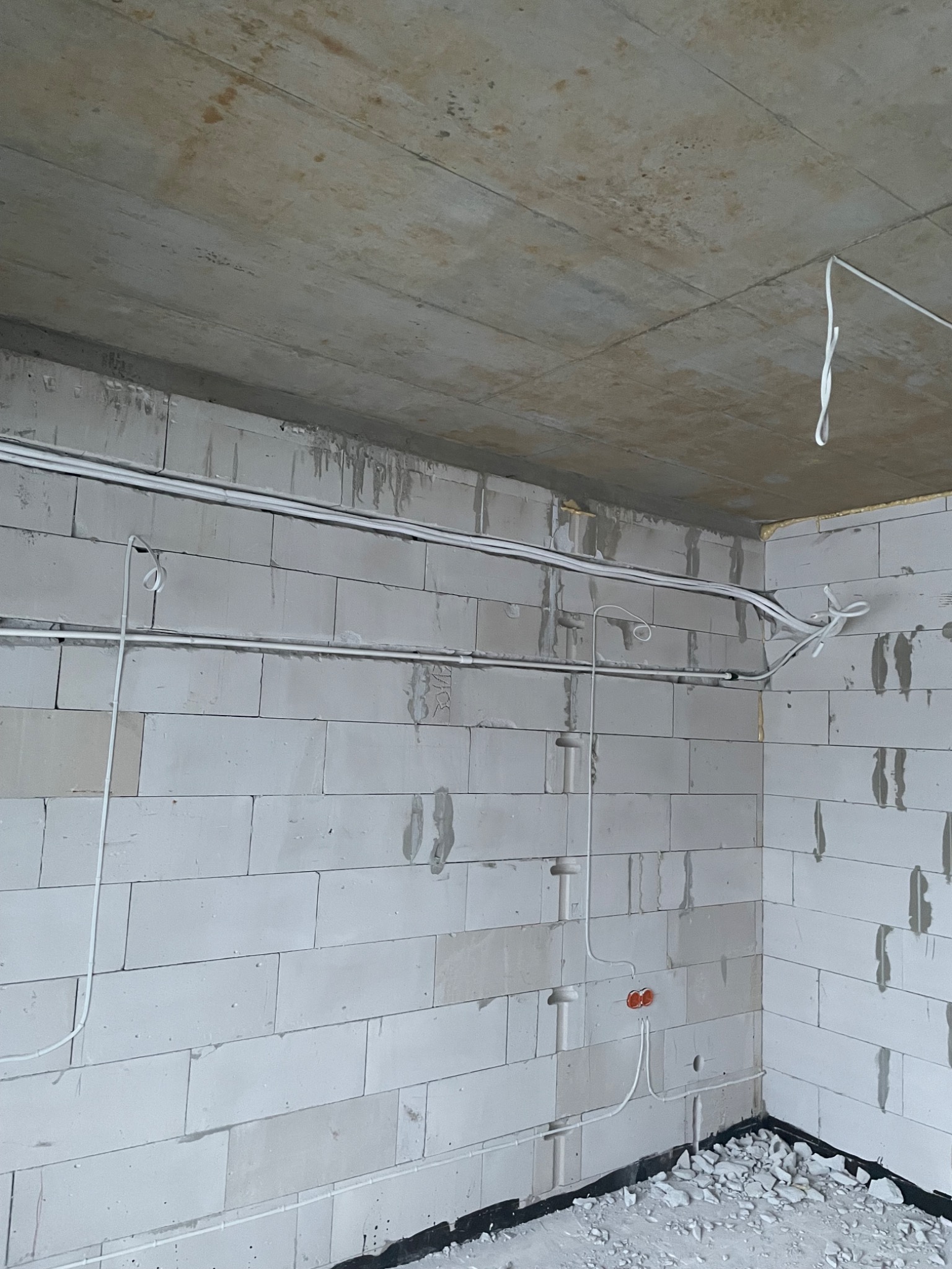 Surowy stan wnętrza budynku w trakcie prac instalacyjnych: widoczne białe ściany z betonu komórkowego, surowy betonowy strop oraz poprowadzone instalacje elektryczne w białych rurkach PCV.