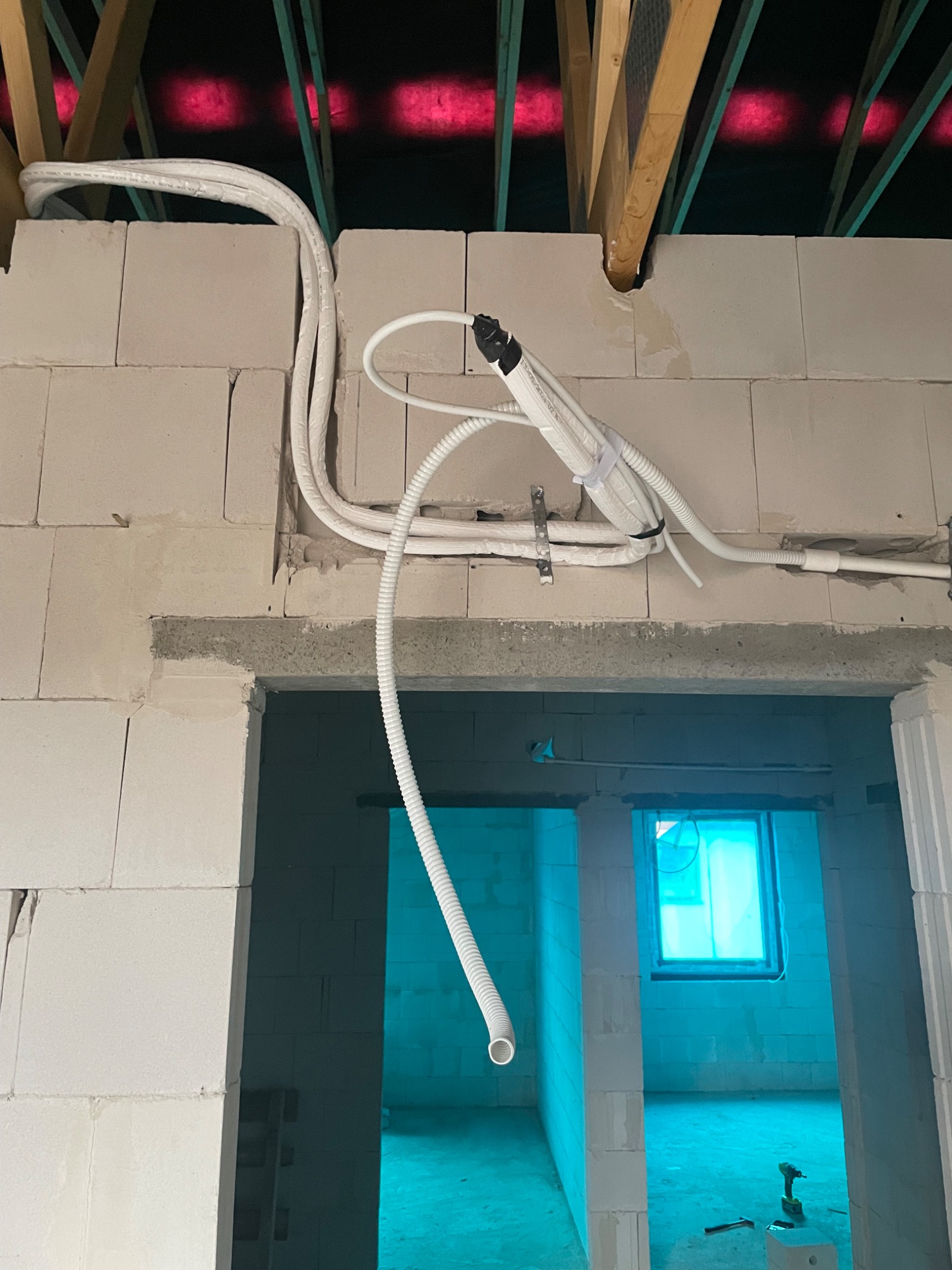 Montaż instalacji wentylacyjnej z rekuperacją w surowym wnętrzu budynku, widoczne białe rury prowadzone pod stropem z drewnianymi belkami, ściany z białych bloczków i nieotynkowane nadproże...