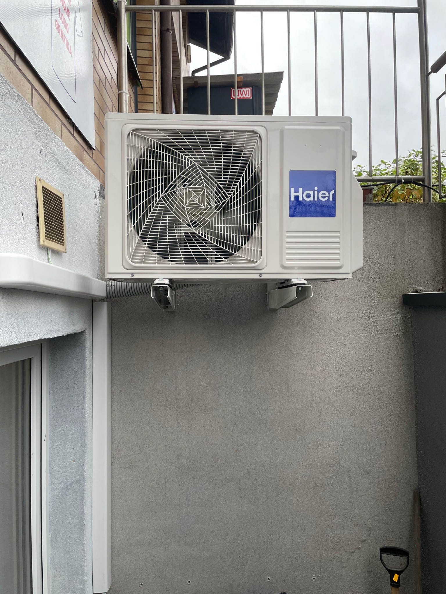 Zewnętrzna jednostka klimatyzacji Haier zamontowana na szarej ścianie budynku, widoczne metalowe wsporniki i peszel odprowadzający skropliny, na górze metalowa balustrada.