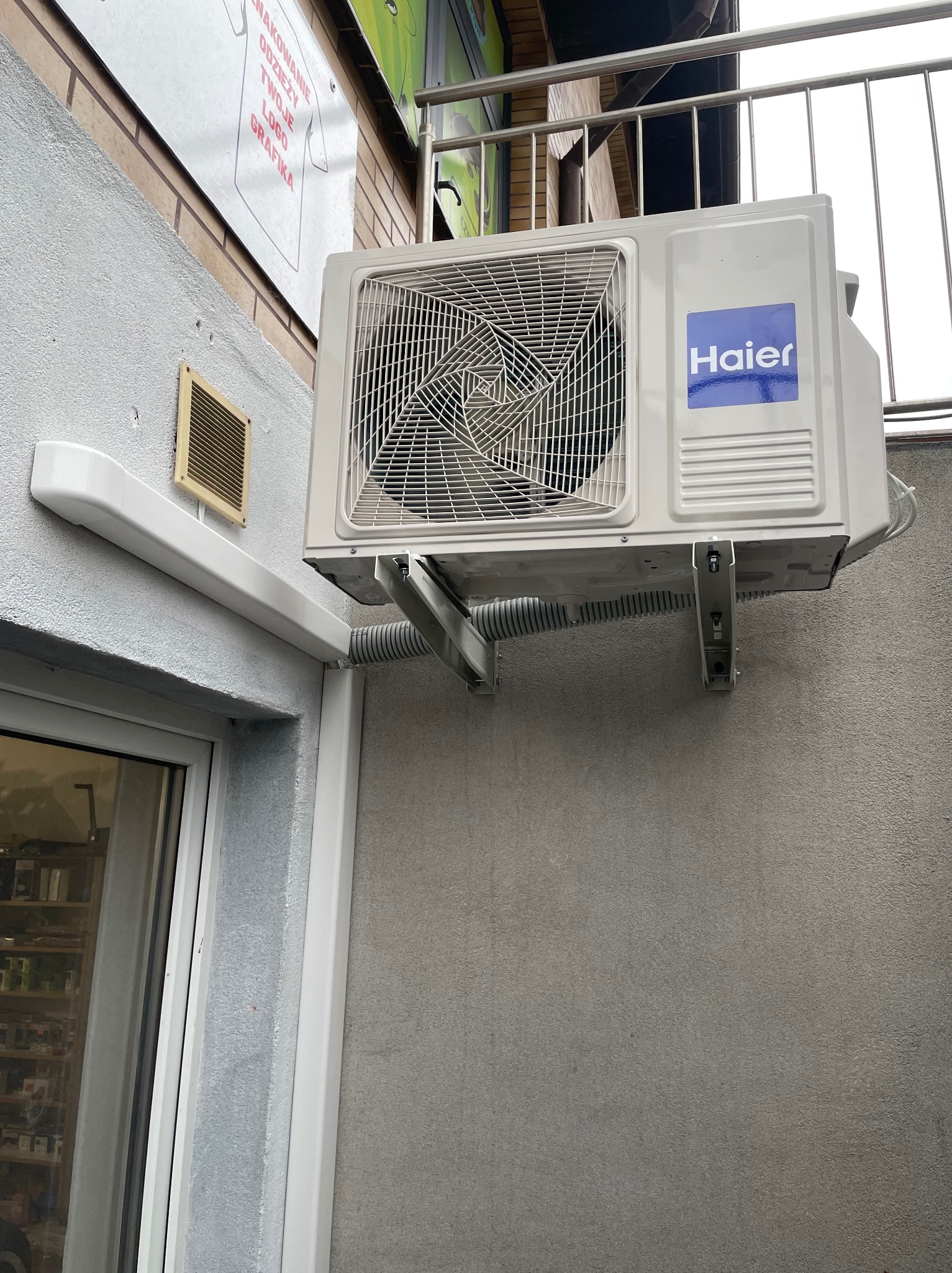 Biała jednostka zewnętrzna klimatyzatora marki Haier zamontowana na szarej ścianie budynku, widoczne metalowe wsporniki i peszel odprowadzający skropliny.