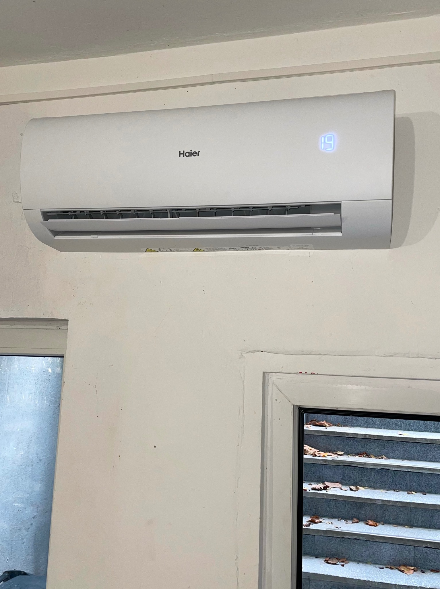 Biały klimatyzator Haier zamontowany na ścianie z widocznym wyświetlaczem temperatury 19 stopni Celsjusza, fragment okna z widokiem na schody.