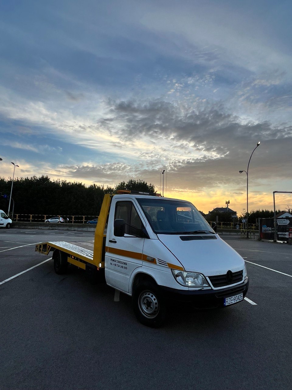 Biała laweta na parkingu o zmierzchu, widoczne oznaczenia 'Pomoc Drogowa' na karoserii, tablica rejestracyjna RZE 9042E, tło stanowi niebo z chmurami i zachodzące słońce.