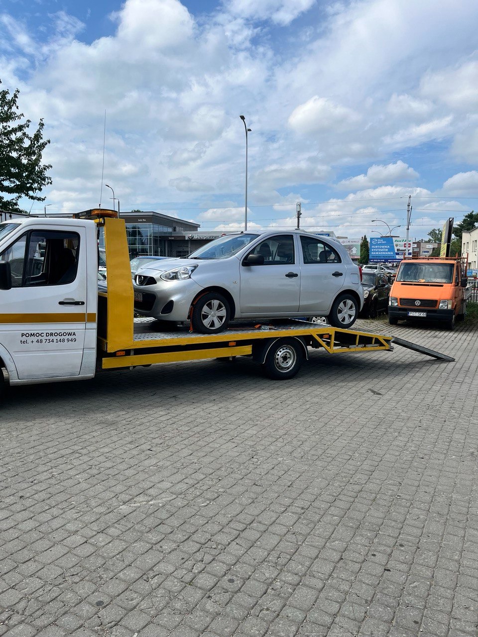 Srebrny samochód osobowy załadowany na żółtą platformę pojazdu pomocy drogowej w Trzebownisku, z widocznym pomarańczowym samochodem serwisowym w tle.