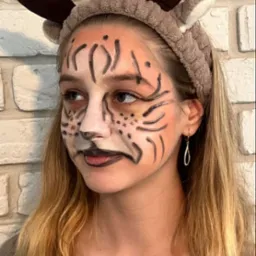 Dziewczyna z pomalowaną twarzą w stylu tygrysa i opaską z rogami renifera na tle ściany z białej cegły.