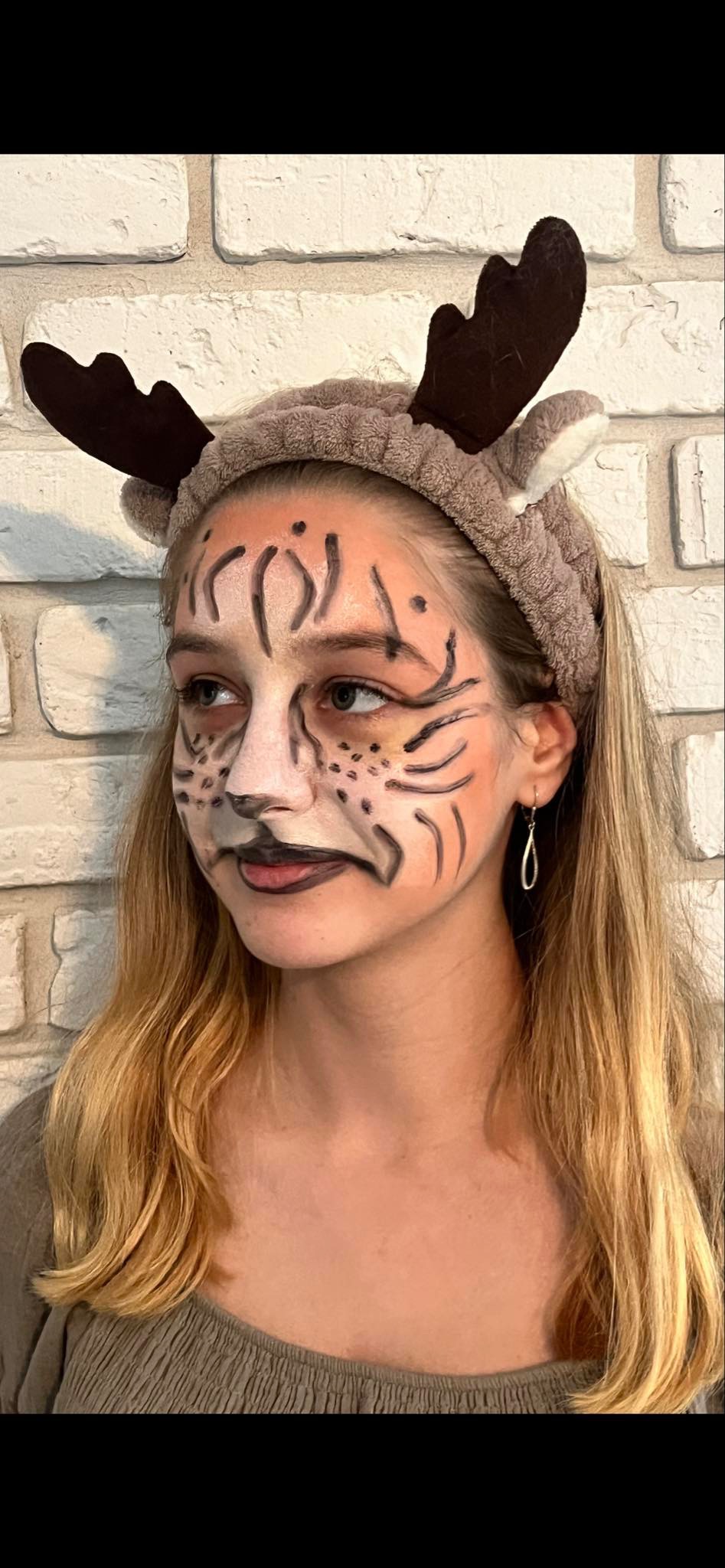 Dziewczyna z pomalowaną twarzą w stylu tygrysa i opaską z rogami renifera na tle ściany z białej cegły.
