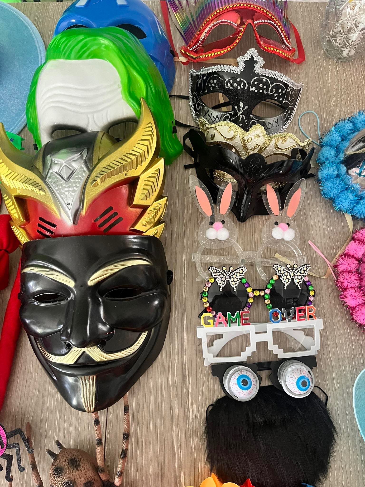 Różnorodne maski karnawałowe i zabawne okulary na drewnianym stole, w tym maska Jokera, Anonymous, królicze uszy i okulary z napisem 'Game Over'.