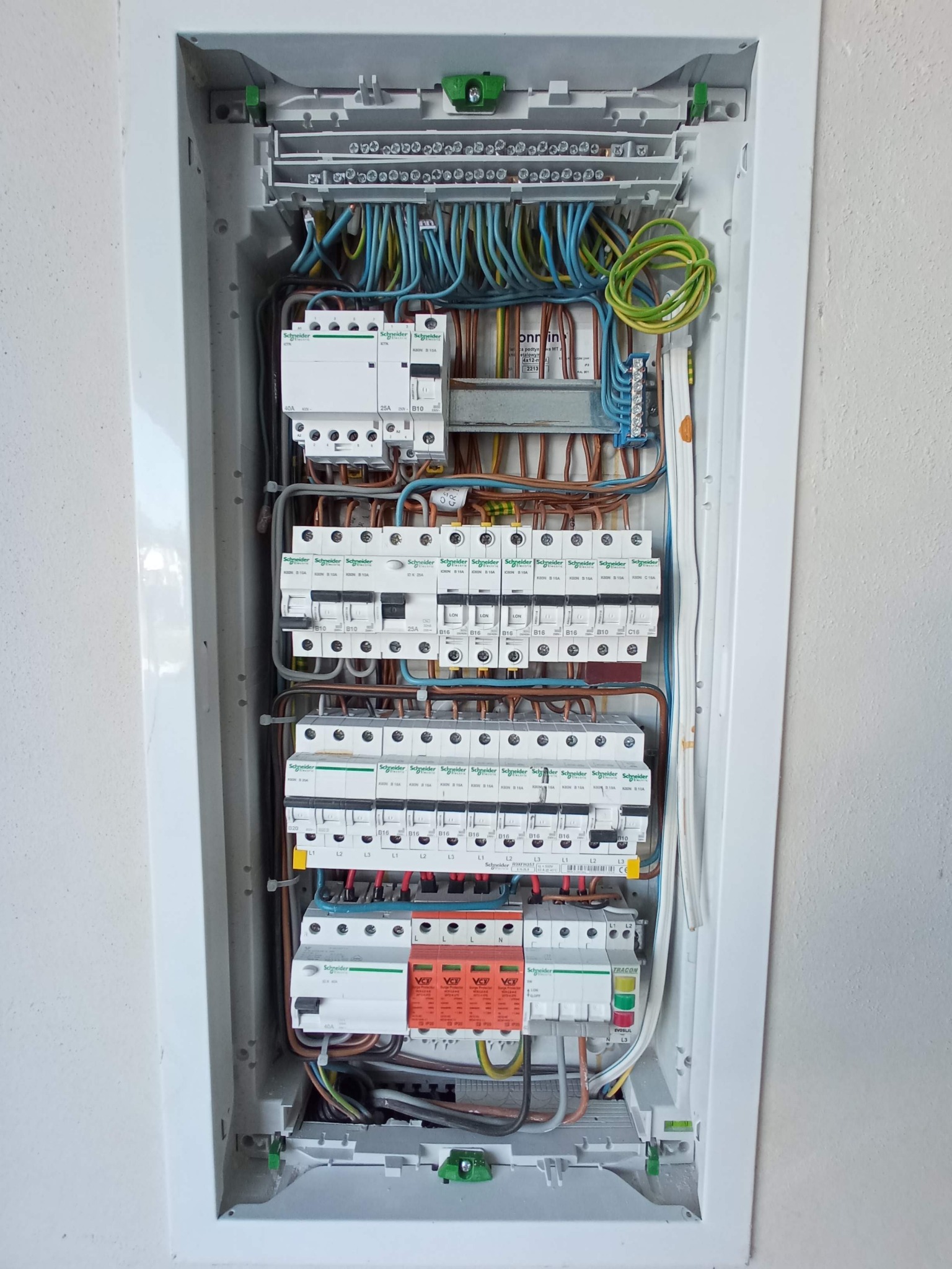 Otwarta skrzynka elektryczna z bezpiecznikami, wyłącznikami i przewodami różnych kolorów, zamontowana na białej ścianie.